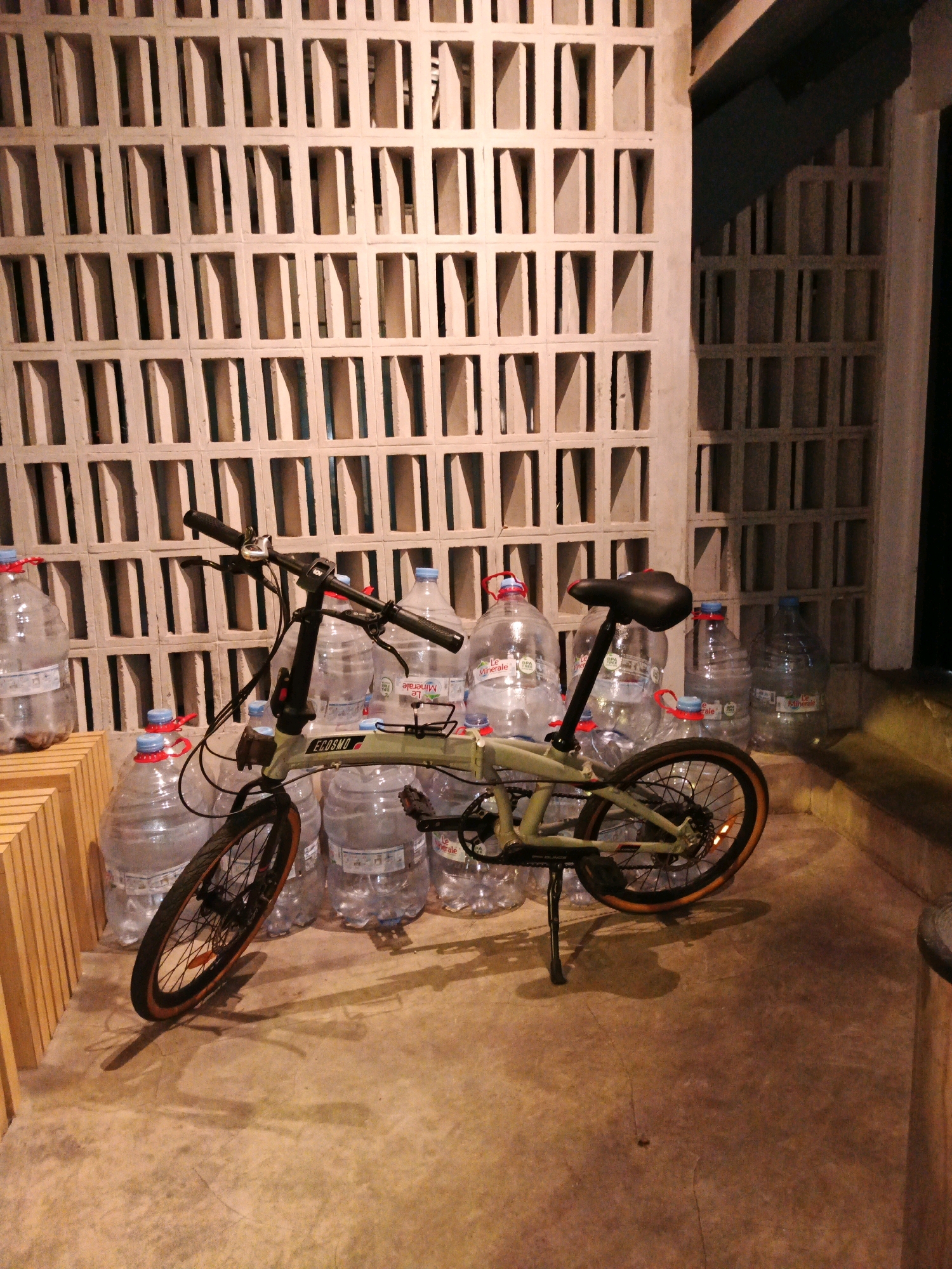 A folding bicycle is positioned in front of a decorative screen wall with rectangular openings. The bicycle is light green with black handlebars and seat. Behind the bicycle, there are several large plastic water bottles with blue and red caps. To the left of the water bottles, there is a stack of wooden slats. The scene is lit by artificial light, casting shadows on the concrete floor. The location is not specified in the image.