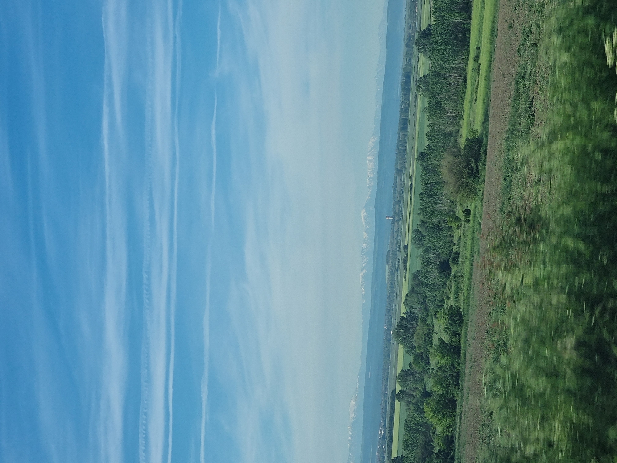 The image presents a landscape scene likely taken from a moving vehicle, with the view skewed due to the angle.
The sky occupies a significant portion of the left side, displaying a light blue color interspersed with streaks of contrails or high cirrus clouds.
The right side features a panoramic view of a terrain that transitions from flat lands to mountains in the distance. It is likely the land surrounding the city of Kozar Belene, Bulgaria. Along the lower margin, a dense green area, potentially a field of crops or grassland, is visible.
Further away, a row of trees delineates a boundary, and beyond that lies a mixture of green fields and possibly some built structures, indicating a settled area. In the far distance, mountains can be seen on the horizon, their peaks possibly snow-capped. The atmospheric conditions appear hazy or slightly overcast, muting the colors and blurring distant details.