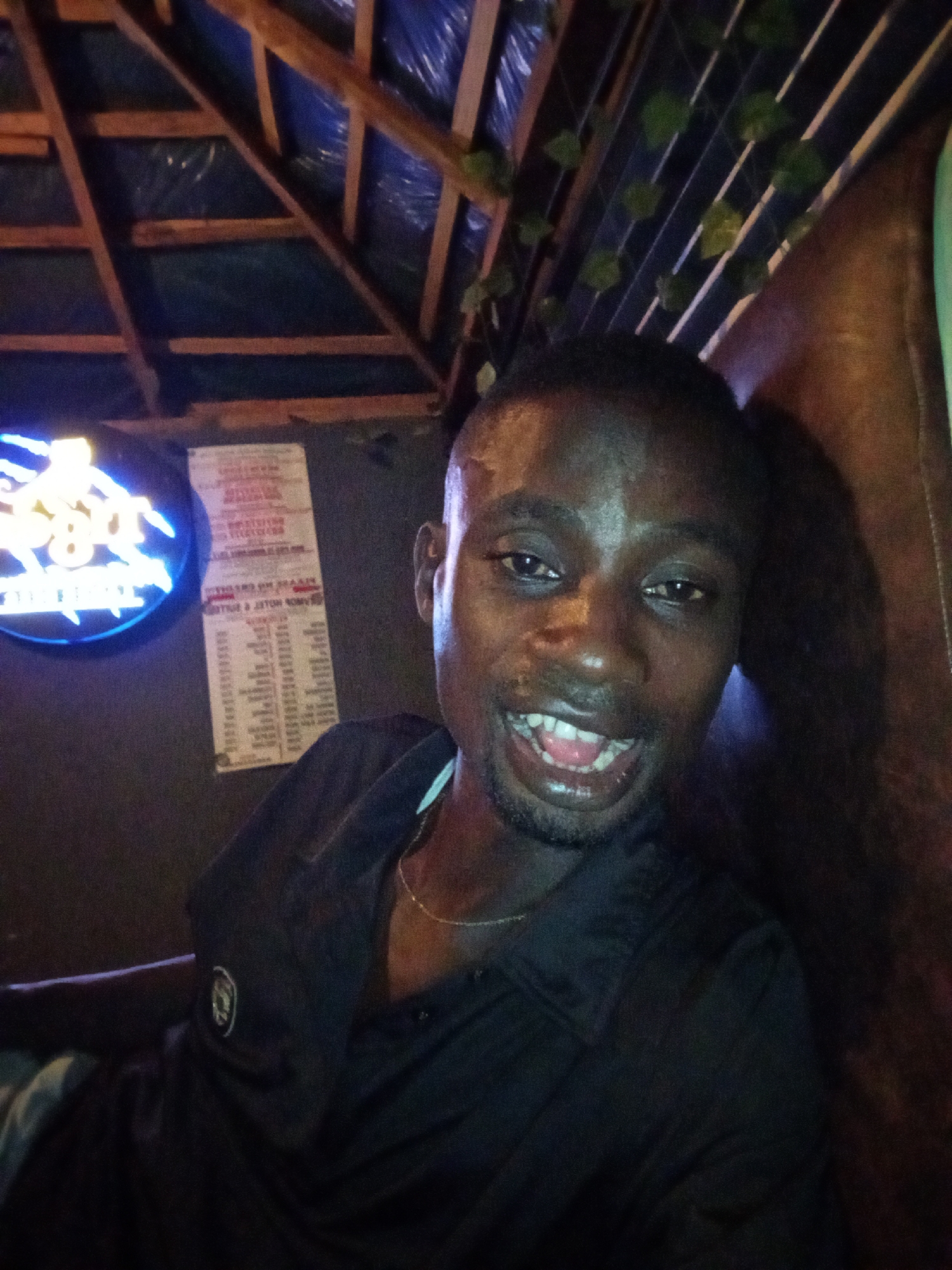 A man with dark skin and short hair is indoors, possibly in a bar or restaurant in Adonkolo, Nigeria. He is smiling and wearing a dark shirt with a logo. A neon sign is visible on the wall behind him, along with a menu or list. The roof appears to be made of wood and plastic sheeting, with some decorative vines. The man is sitting on a leather couch.