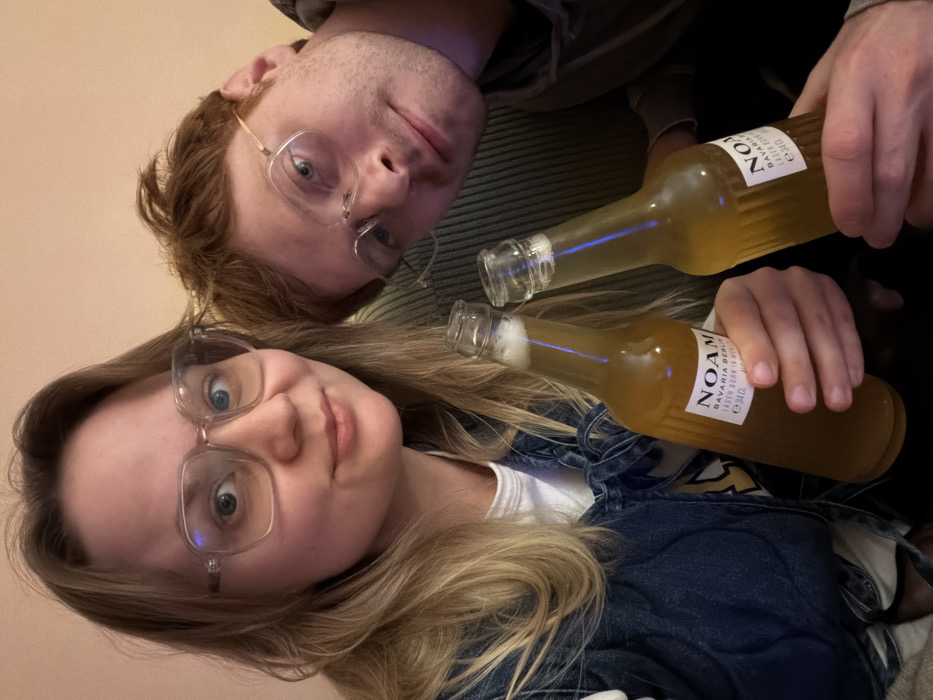 The image captures two individuals, a female and a male, in a close-up indoor shot. The female, positioned on the left, has long blonde hair, blue eyes, and wears large clear-framed eyeglasses, a white t-shirt, and a blue denim jacket. She holds a clear glass bottle labeled "NOAM BAVARIA BERLIN LAGER BORN IN MUNICH e 34 cl" in her left hand. The male, on the right and slightly behind her, has reddish-brown hair, blue eyes, and wears wire-frame eyeglasses and a dark hooded top. He holds an identical "NOAM" beer bottle in his right hand. Both individuals are looking directly at the camera with neutral to slightly smiling expressions. The bottles contain a light amber liquid with some foam visible at the top. The background is an indistinct, plain, light-colored wall, suggesting an interior setting under artificial light. The specific location within the city of Tel-Aviv, Israel, cannot be determined from the visual evidence in the image.