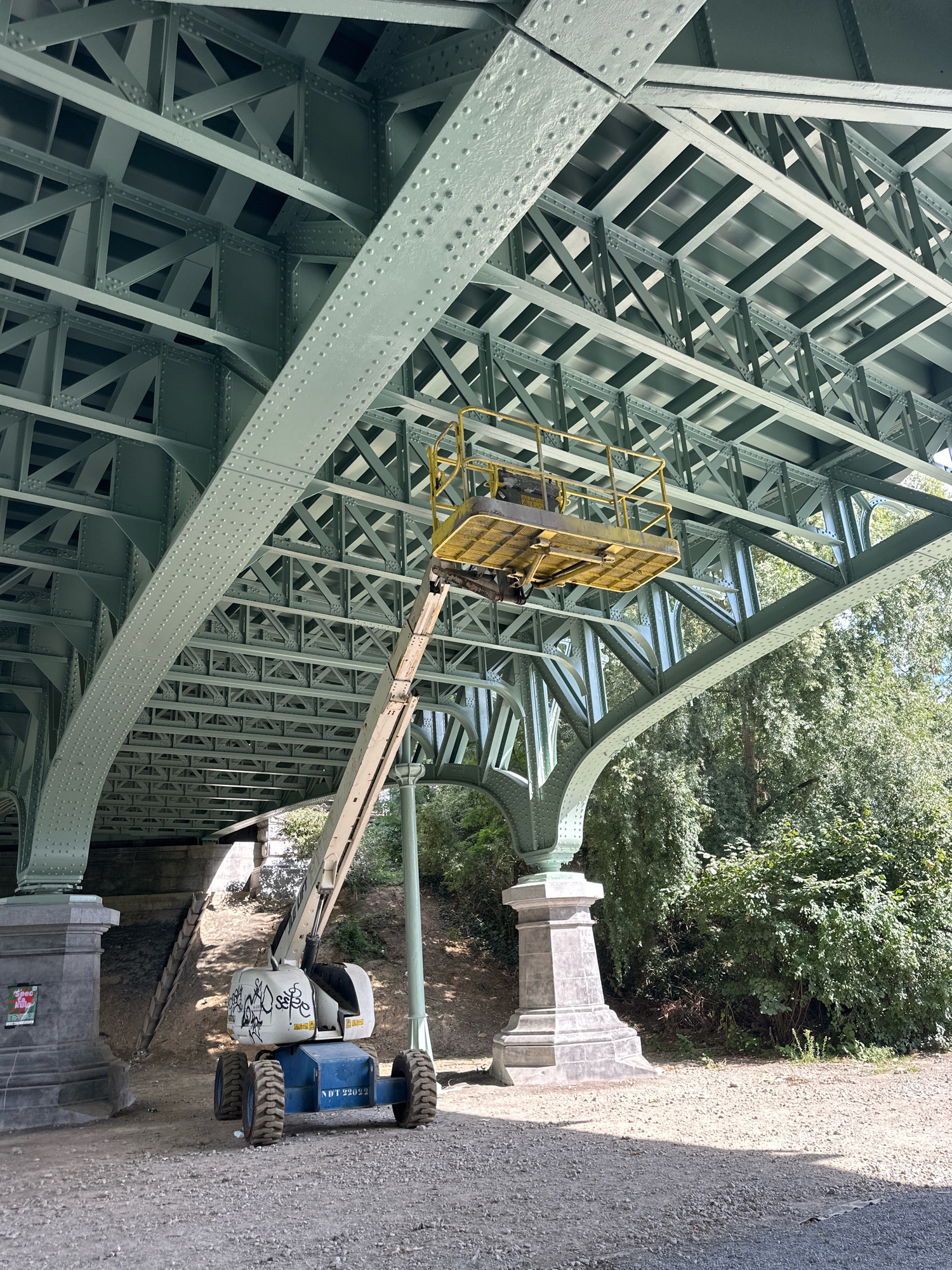 Underneath a large, green, riveted metal bridge structure, a white and blue aerial lift platform with a yellow basket is positioned. The lift's arm extends upwards towards the bridge's underside, where structural beams and supports are visible. The ground beneath the bridge is a gravel and dirt path, with some vegetation and trees in the background. The overall scene suggests construction or maintenance work is taking place on the bridge. The location is identified as Molenbeek-Saint-Jean - Sint-Jans-Molenbeek, Belgium.