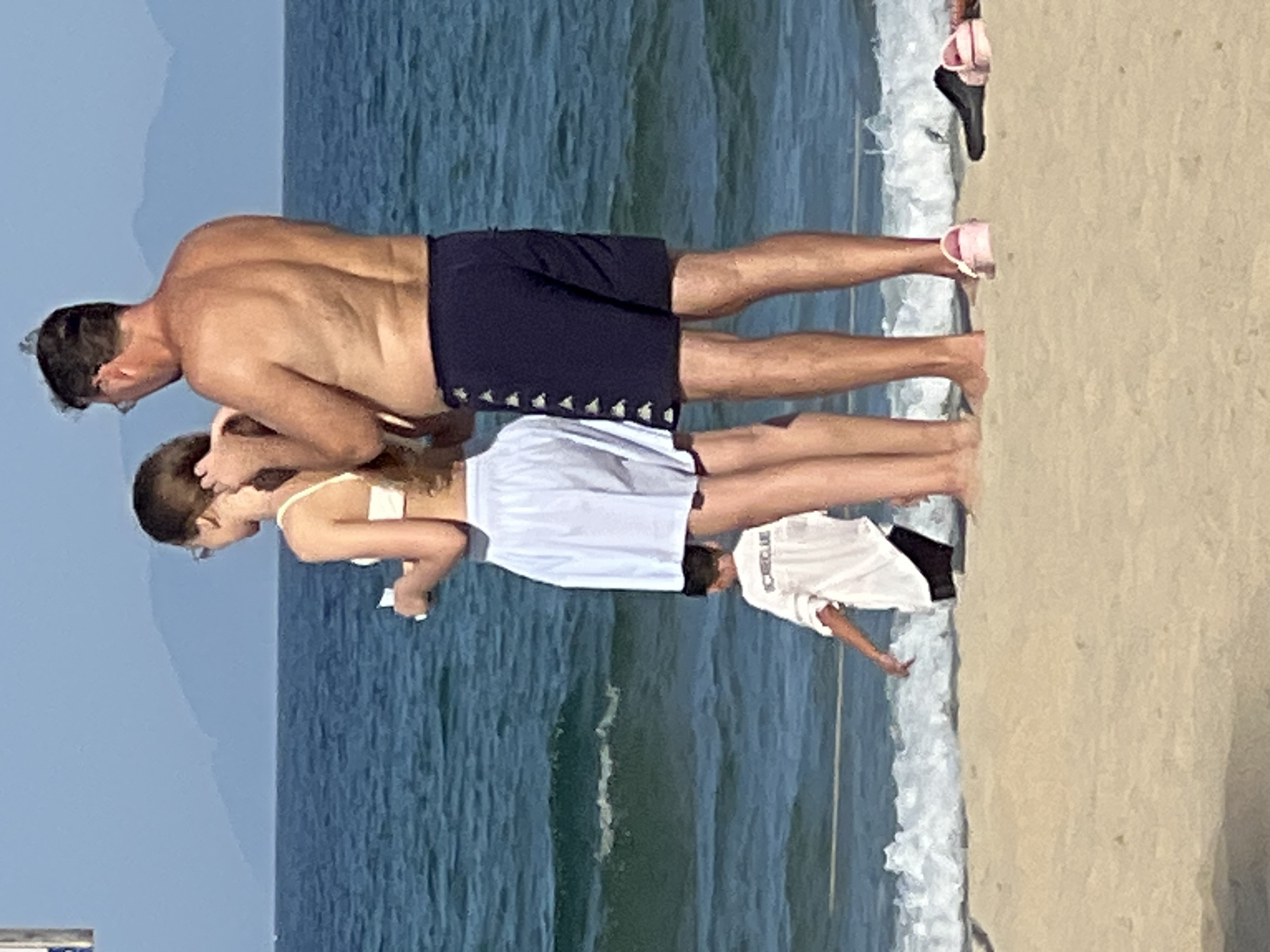 A man and a girl are standing on a sandy beach with the ocean in the background. The man, who is shirtless and wearing dark blue swim trunks, is leaning over. The girl is wearing a white tank top and a white skirt. She is looking down at something in her hand. To the right, another person, possibly a boy, is visible from the waist down, wearing a white t-shirt and dark shorts. The sun appears to be shining, casting shadows, suggesting it is daytime. The water is blue with gentle waves. The scene is set in Đà Nẵng, Vietnam.