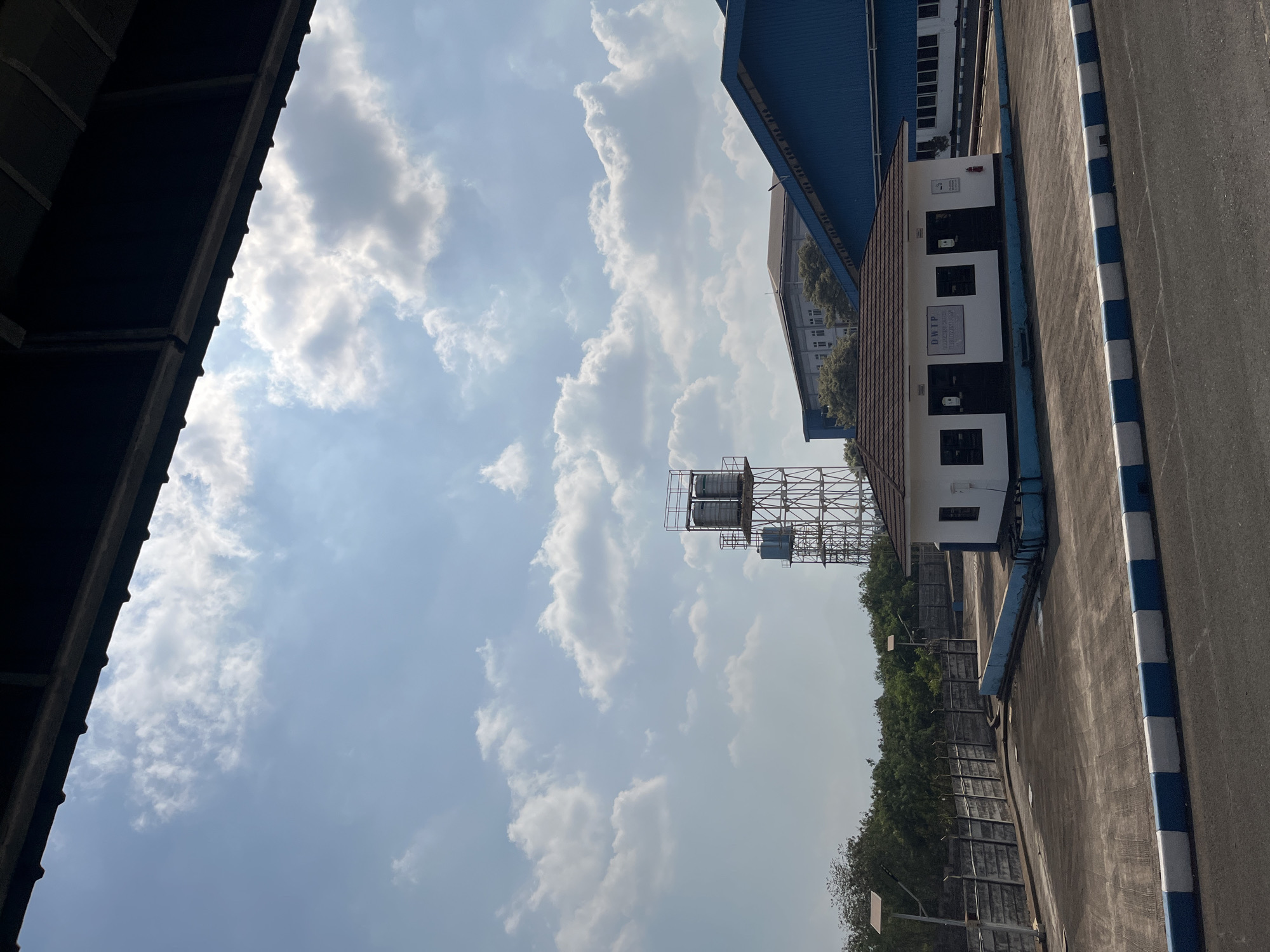 The image depicts an outdoor scene on a bright, partly cloudy day, likely in the city of Pelang, Indonesia, given the context of the prompt. In the foreground, a paved area with a distinct blue and white striped curb is visible, suggesting a road or parking lot. Beyond this, a small, white building with a brown tiled roof stands prominently. The building has two dark doors and two dark windows on its front facade, and text that reads "DWIP" is visible above the doors. To the left of this building, a grey concrete wall topped with barbed wire runs along the perimeter of the property, with solar-powered lights mounted on poles along its length. Behind the wall, lush green trees form a dense backdrop.
In the mid-ground, a tall, metal water tower structure is visible, supporting at least two large cylindrical tanks, with a smaller blue tank below them. Further back, the roofs of larger industrial buildings are present, one appearing to be a blue, pitched roof structure characteristic of a factory or warehouse. The sky above is a clear blue, dotted with fluffy white clouds, and sunlight illuminates the scene. There are no people or animals visible in the frame.