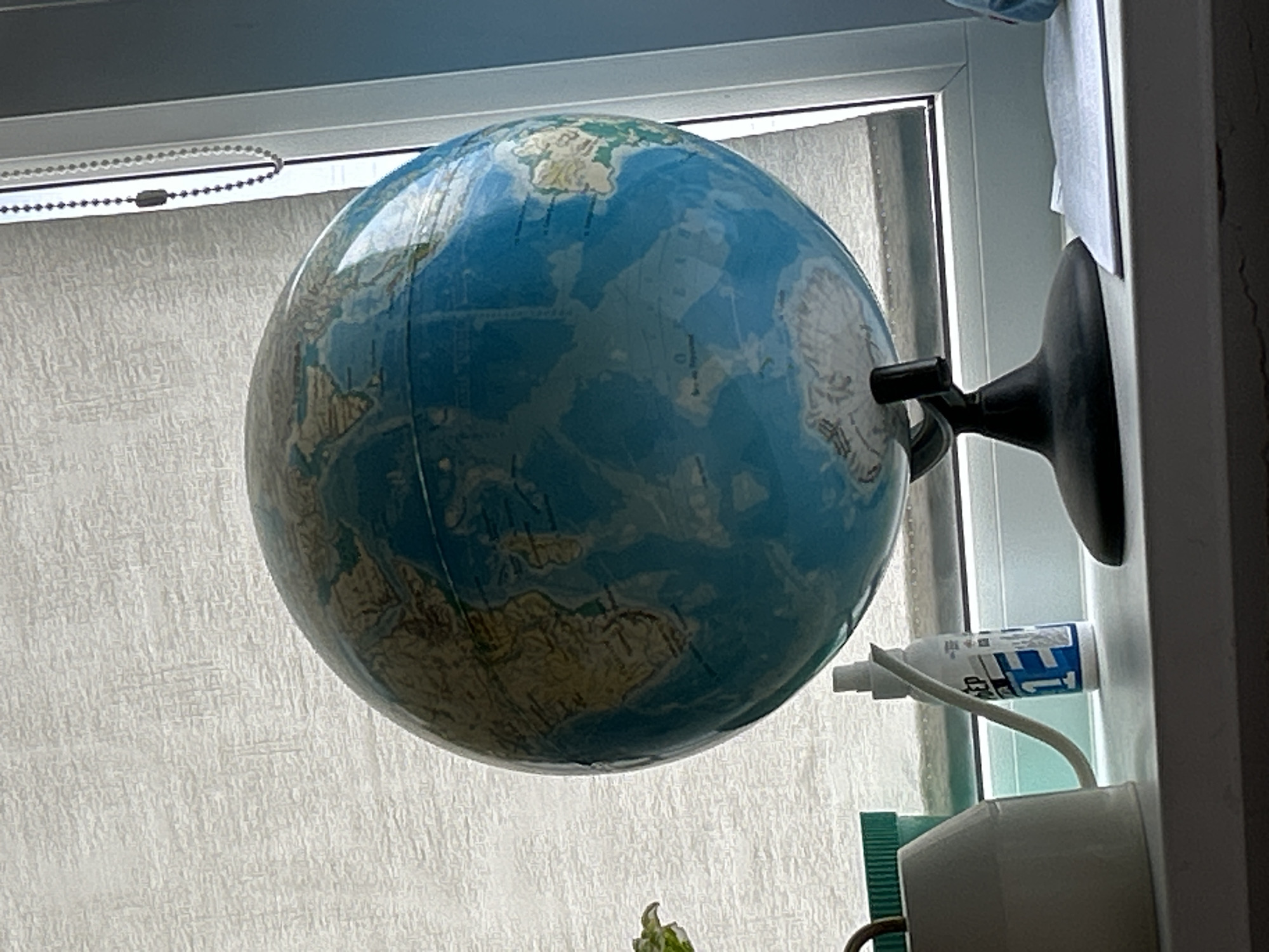 A static shot captures a physical globe positioned in front of a window, likely in Chaikovsky, Russia. The globe features light blue oceans and tan/green landmasses, with North America, Greenland, parts of Eurasia, and Antarctica prominently visible. Its surface appears to have geographical contours, suggesting it might be a relief globe. It is mounted on a black plastic arm and base, which is attached to the white window frame or an adjacent structure on the right side of the image.
Behind the globe, the window is covered by white, textured blinds or curtains, which are illuminated by bright daylight, indicating the photo was taken during the day. A beaded metallic chain, typically used for operating blinds, hangs in the upper left portion of the window.
To the right of the globe, resting on a white ledge or window sill, is a white spray bottle with a blue and white label. Partially visible text on the label includes "T1". Below this, a white electrical device, possibly a charger or air freshener, with a white cable, is also present. Faint geographical names are discernible on the globe's surface, but are not clearly legible. No specific activity is taking place; the scene is static.
