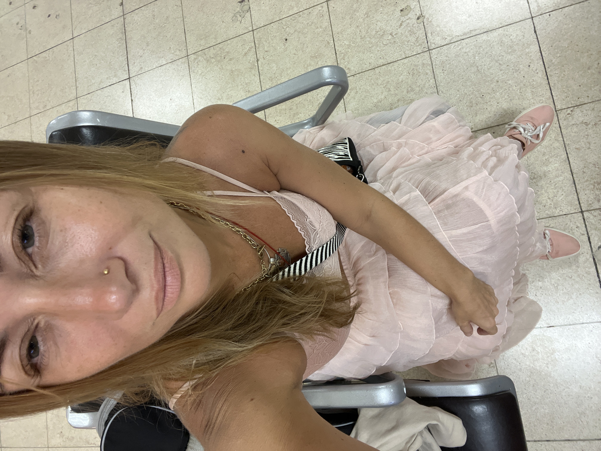 The image is a selfie of a blonde woman with fair skin and light freckles, sitting in what appears to be a waiting area. She is wearing a light pink, ruffled dress with spaghetti straps, a lace bralette visible underneath, and pink sneakers. She has a gold nose stud and is wearing a layered necklace. She is holding one hand near her lap, with her other arm resting on the armrest of a gray and black chair. The environment appears to be a tiled floor and seating area, possibly in an airport or public waiting room, consistent with locations in Katunayake, Sri Lanka, which is known for its proximity to Bandaranaike International Airport.