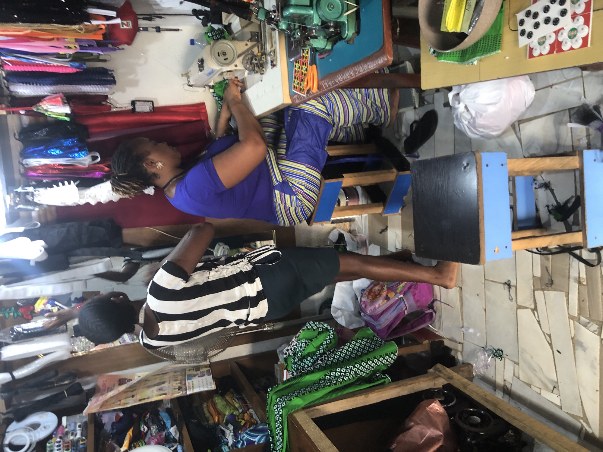 This image depicts a tailor's shop in Ado, Nigeria, with two women actively engaged in sewing. One woman, wearing a black and white striped top and a dark skirt, stands by a large oscillating fan, her back to the viewer. The other woman, seated at a sewing machine, is wearing a purple top and horizontally striped pants. She is focused on her work, with a sewing machine and various fabrics spread out on the table in front of her. The shop appears cluttered, with shelves packed with colorful fabrics, rolls of material, and sewing supplies. A small stool with a blue frame and a black seat is positioned near the woman at the sewing machine. Various bags and items are scattered on the floor, indicating a busy working environment. The walls are adorned with hanging fabrics, ribbons, and decorative items. The overall impression is that of a functional and active tailoring business.