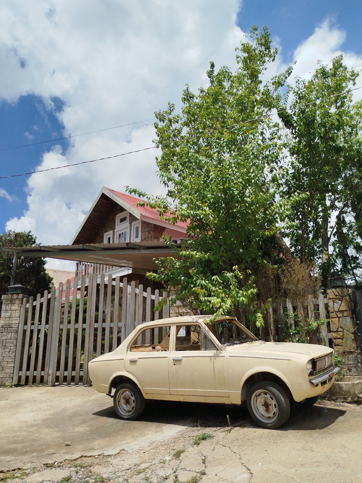 An abandoned, faded yellow sedan sits parked on a cracked, dusty road in front of a weathered wooden fence. Behind the fence, a two-story house with a red, corrugated metal roof and stone exterior is visible. Lush green trees with dense foliage grow in front of the house. The sky above is a vibrant blue, dotted with fluffy white clouds. The scene suggests a rural or suburban setting, possibly in the city of Xuân Trường - Đà Lạt, Vietnam, with a focus on the neglected vehicle and its surroundings. There are no people present in the image.