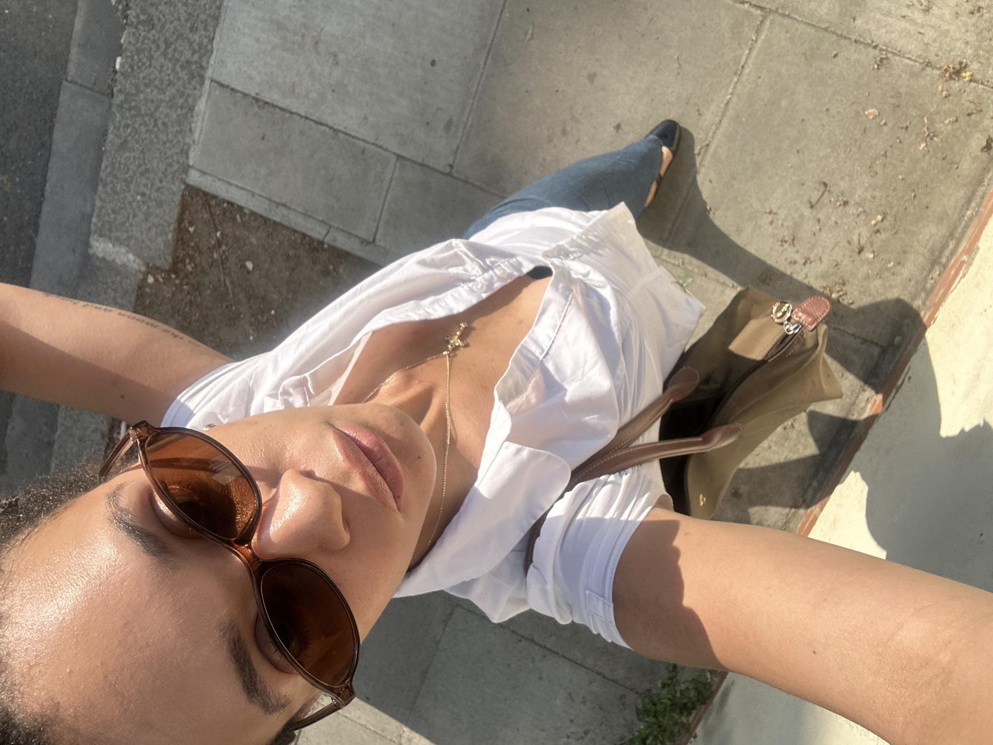 A downward-angled, wide-shot perspective shows a person standing on a sunlit sidewalk, likely in a city context. The individual wears round, brown-framed sunglasses, a white button-down shirt, blue jeans, and black flats. A brown purse is positioned near their leg. The sidewalk is composed of gray slabs. A tattoo is visible on the person's arm.