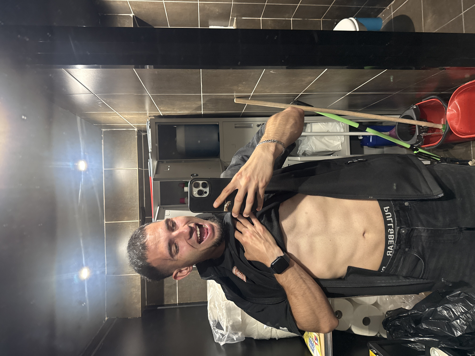 A man is taking a selfie in a bathroom. He is shirtless, wearing black jeans and a black vest, and holding up his phone to capture the image. The man is grinning with his mouth open. The bathroom has brown tiled walls, and there is a mirror reflecting the scene. Behind the man, there's a rack with toilet paper, cleaning supplies, and a bucket. The exact location in Shumen, Bulgaria, cannot be determined from the image.