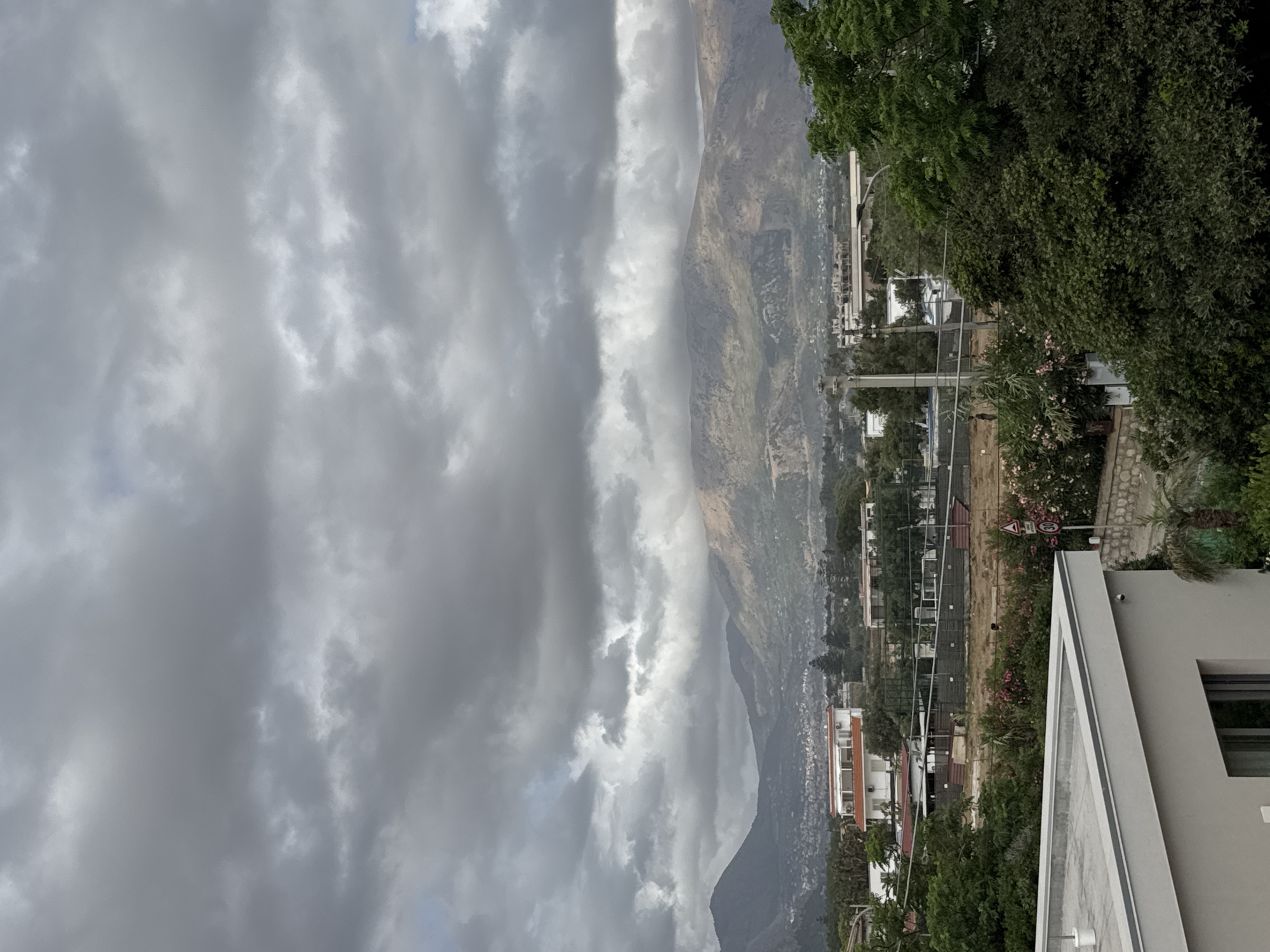 The image depicts a cloudy, overcast sky over a suburban or semi-rural landscape in Capaci, Italy. In the foreground, a modern, light-colored building with a flat roof is partially visible on the left. Beyond this building, a dense arrangement of trees and vegetation is present. Further into the middle ground, several buildings are visible, including a multi-story structure with a red-tiled roof and other smaller houses and structures, some with what appear to be sports facilities or fenced-off areas. In the background, a range of mountains dominates the horizon, their slopes covered in vegetation and rocky outcrops. A town or city is nestled at the base of the mountains. The overall atmosphere is subdued due to the heavy cloud cover. No people are clearly discernible in the image.