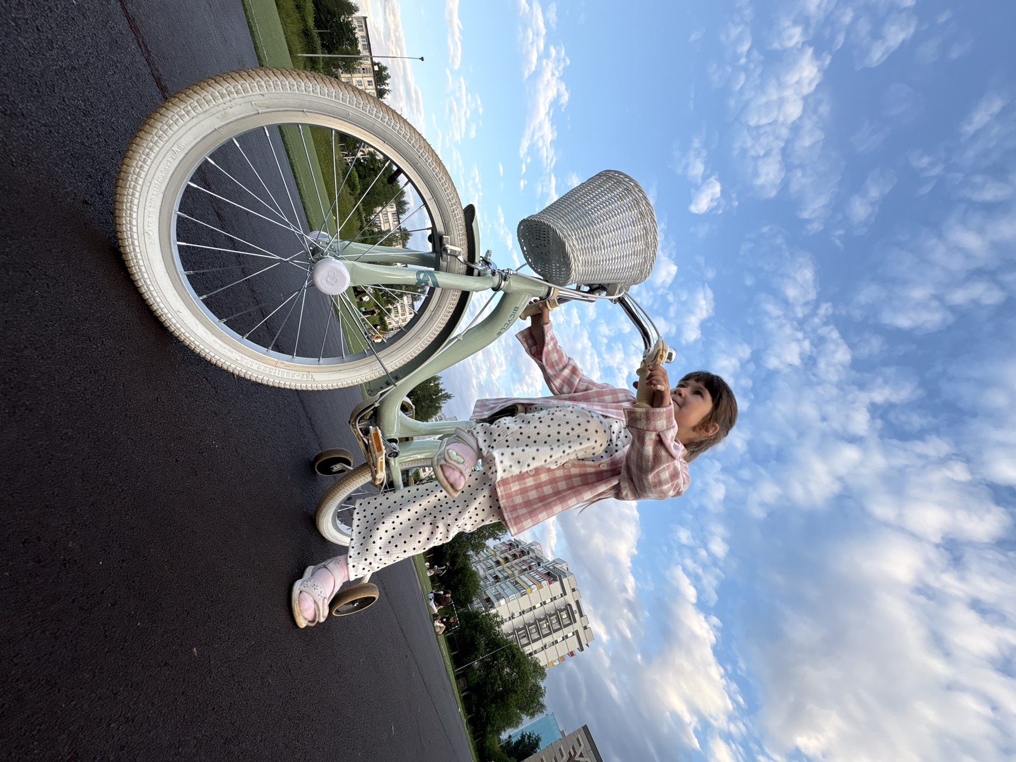A young girl, dressed in a pink gingham jacket and white pants with black polka dots, rides a light green bicycle with training wheels. She is wearing pink sandals. The bicycle has a white wicker basket on the handlebars and a large white tire. The girl is looking up and to the right, with a curious expression. The image is taken from a low angle, looking up towards the sky, which is filled with scattered white clouds against a bright blue sky. In the background, there are trees, a green sports field, and modern apartment buildings with colorful balconies. The scene appears to be set in the late afternoon or early evening, with soft lighting. The ground is asphalt. Visible text on the bicycle tire reads "INFLATE TO 20-30 PSI (1.4-2.0 BAR) 65-120 kPa". The location context is Chaikovsky, Russia.