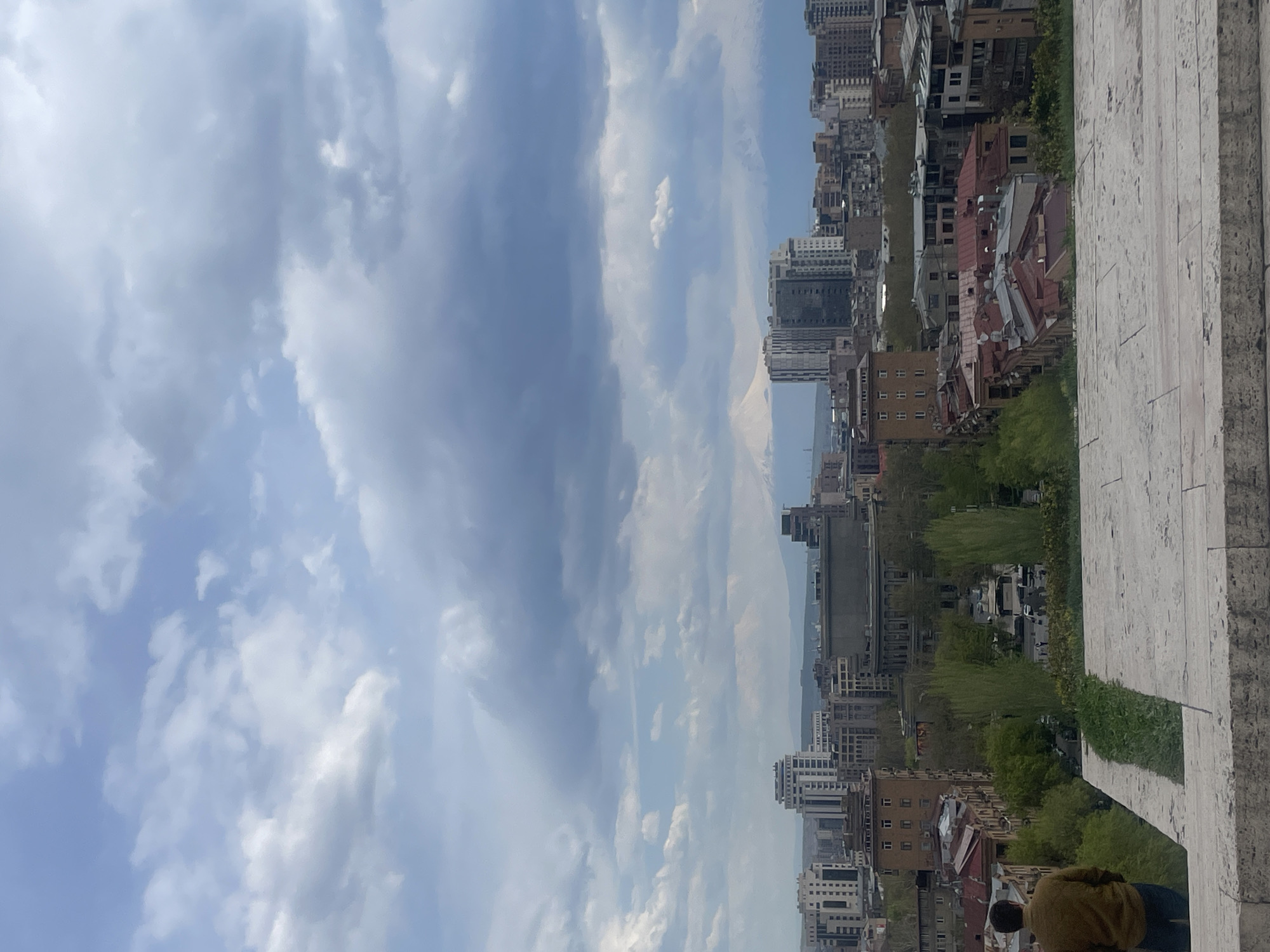 The image presents a view of Yerevan, Armenia, from an elevated position, possibly from the Cascade complex. The foreground shows a stone structure, suggesting a vantage point within the Cascade itself. The cityscape stretches out below, featuring a mix of residential buildings, commercial structures, and green spaces with trees. The architecture is varied, with buildings of different heights and styles. In the distance, a mountain range is visible through a break in the urban landscape. The sky is mostly covered with clouds. There are at least two people in the foreground.