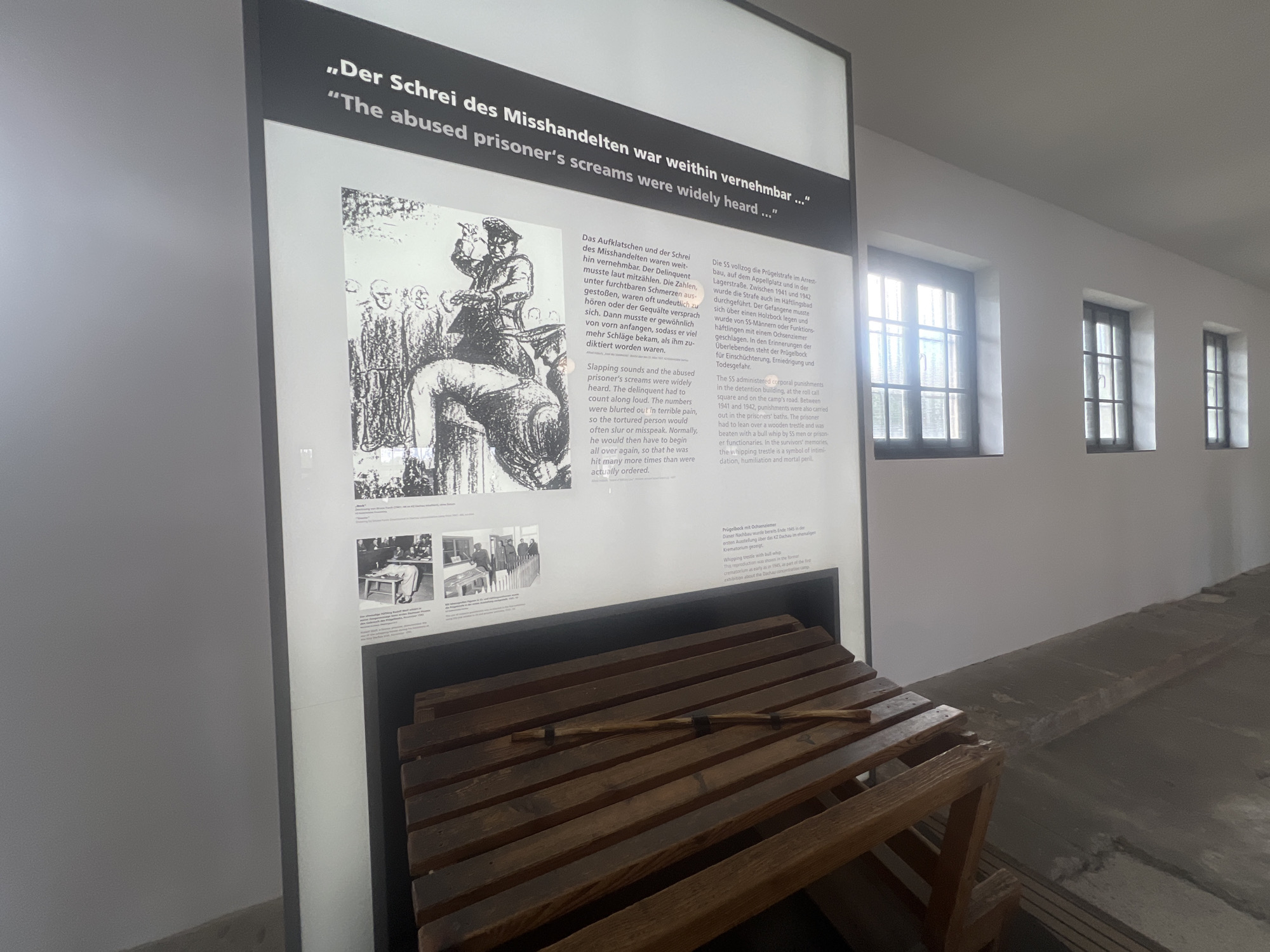 The image depicts an exhibit within the Dachau Concentration Camp Memorial Site in Dachau, Germany. The primary focus is a large, vertical display board with text in German and English. The German text at the top reads, "„Der Schrei des Misshandelten war weithin vernehmbar...“" and the English translation below is, "„The abused prisoner's screams were widely heard...“".
To the left of the text is a black and white illustration depicting a historical scene of punishment, showing a figure standing over a person on the ground, surrounded by onlookers. Below this illustration are two smaller black and white photographs. The top photograph shows a line of people in what appears to be a roll call or assembly area. The bottom photograph shows an individual bending over a table with other people in the background.
In the foreground, a wooden exhibit is placed. It consists of a sloped wooden structure made of slats, with a thin wooden stick, possibly a whip or a baton, resting horizontally across it. This exhibit appears to be designed to represent an instrument of punishment or an object related to the depicted historical events.
The background of the image shows a section of a building with white walls and three large windows with dark frames. The windows are divided into multiple panes, and light is filtering through them, creating reflections on the wall. The floor appears to be a light gray concrete. The overall environment suggests an indoor exhibit space within a historical memorial.