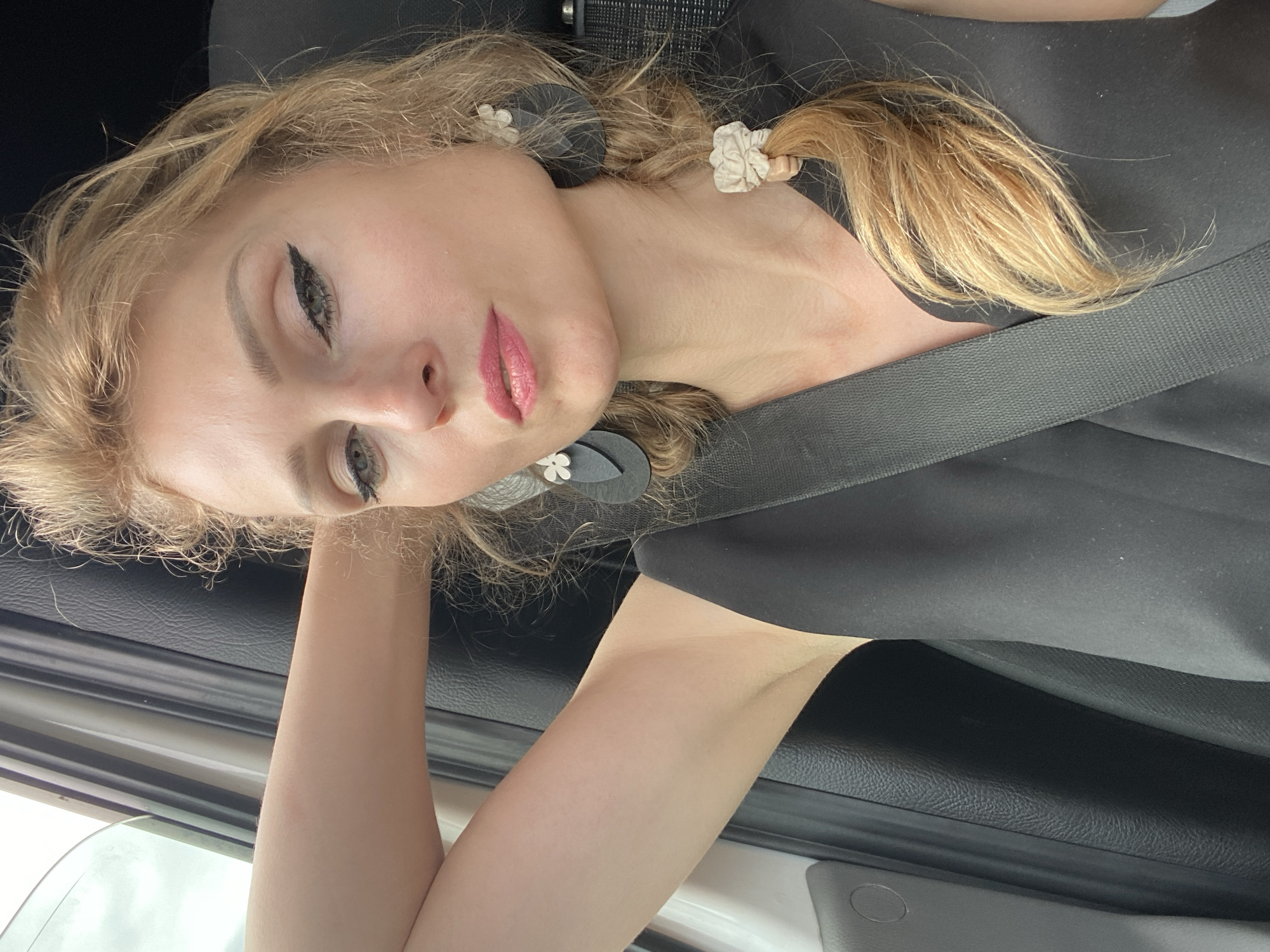 A blonde woman with braided hair and makeup is in the passenger seat of a car. Her left arm is raised behind her head, resting on the headrest. She is wearing a black sleeveless dress and a seatbelt. The car interior is visible, with a black leather seat and a white door panel. The background outside the window is blurred and appears to be a daytime scene, possibly in a rural or suburban area. The specific location in Валожынскі сельскі Савет, Belarus, cannot be determined from the image alone.