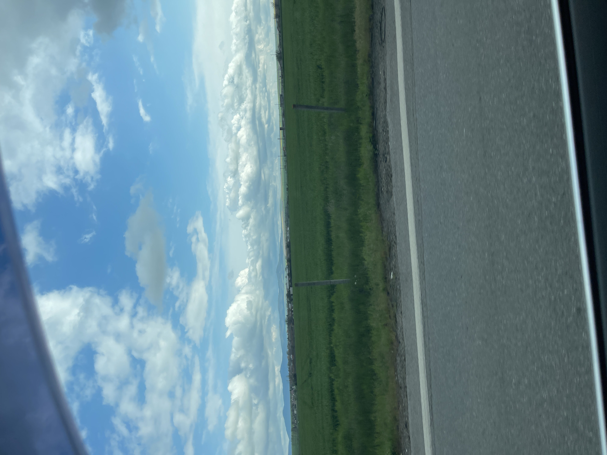 The image shows a view from a vehicle, likely a car, traveling on a road in Vodenichane, Bulgaria. To the right, the gray asphalt road is visible with a white line marking the edge. A green field stretches along the other side of the road with scattered posts or markers extending into the distance. The sky fills the upper left portion of the frame and is filled with blue sky and white clouds. A partial view of the vehicle's window frame is seen on the left and right edges.