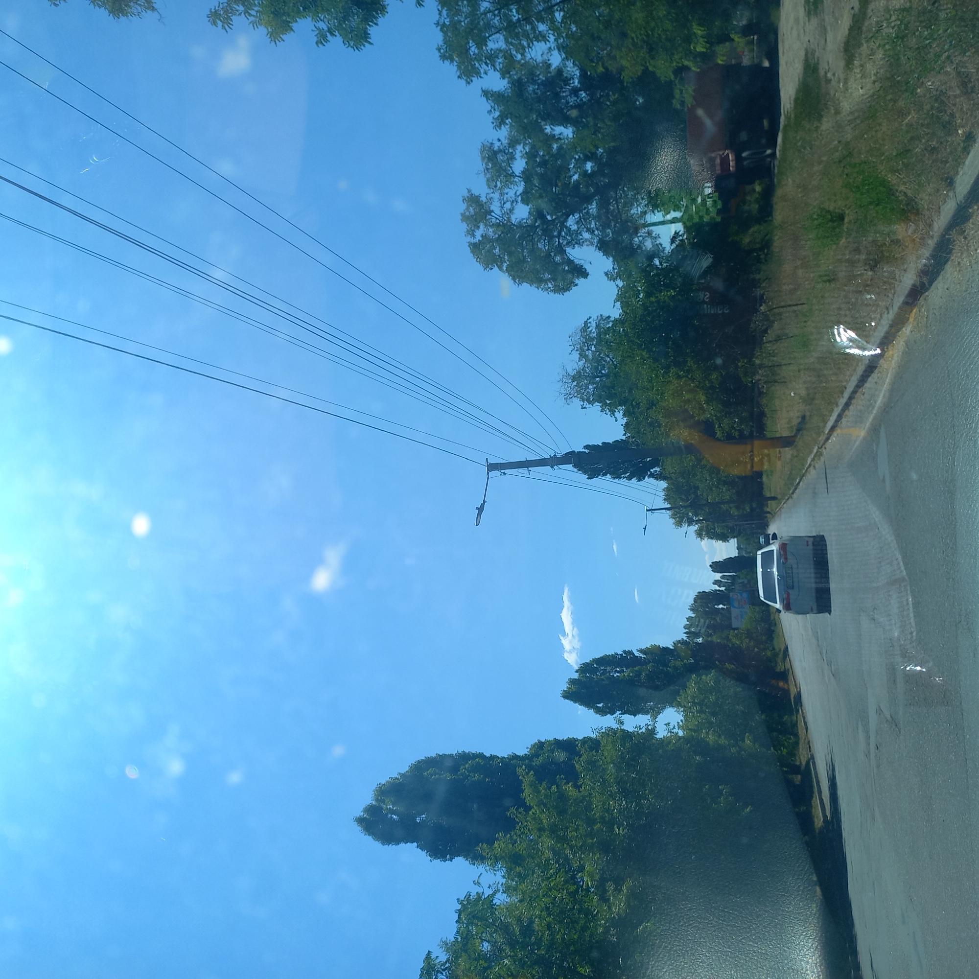 The image depicts a sunny day in Kropyvnytskyi, Ukraine, taken from a vehicle traveling on a road. The road has two lanes and a visible curb on the right side. Trees line both sides of the road, with tall, slender trees interspersed with broader, leafy ones. Power lines stretch across the blue sky, supported by utility poles, one of which has a street light attached. A white sedan is visible in front, driving in the same direction. In the distance, on the right side of the road, a large truck is parked or moving slowly. The overall scene suggests a suburban or semi-rural setting within the city.