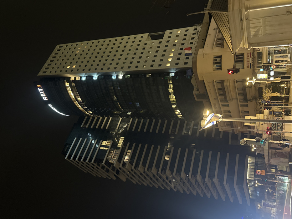 The image depicts an urban street scene at night, centered on Rothschild Boulevard in Tel Aviv, Israel.
In the background, two prominent modern skyscrapers dominate the skyline. The skyscraper on the center-right is tall with a dark, curved glass facade, featuring numerous illuminated windows displaying yellow and blue light. At its apex, an illuminated sign reads "REIT 1". To its left, another tall, dark-facaded skyscraper is visible, characterized by multiple parallel horizontal white architectural bands or balconies, with some interior lights visible through its windows.
In the mid-ground, below the skyscrapers, stands an older, lighter-colored multi-story building, likely of Bauhaus or International Style architecture. This building has multiple windows and a sign on its facade that reads "INSTITUT FRANCAIS". On the roof of this building, two small flags are visible: one red, one blue.
The foreground shows street-level elements. A tall pole with an Israeli flag attached is visible to the left of the Institut Français building. Multiple streetlights illuminate the area, and traffic lights display both red and green signals. Various street signs with Hebrew text are present. On the far right, part of a light-colored, small building or kiosk with a gabled roof is visible. Indistinct figures of people can be observed on the sidewalk in the lower left and center, illuminated by streetlights. The overall scene is lit by artificial urban lighting against a dark night sky.