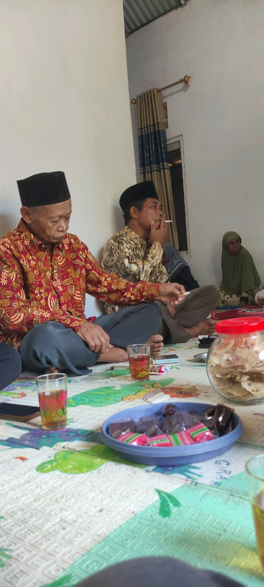 The image depicts an indoor gathering with several people sitting on the floor, which is covered by a colorful mat. In the foreground, a glass of tea and a bowl of assorted candies are visible. To the left, an elderly man wearing a black cap and a red patterned shirt is seated. To his right, another man with a black cap is smoking a cigarette. Further back and to the right, a woman in a green hijab is also present. The background shows a white wall with a curtain and a window. The overall scene suggests a casual social event or a family gathering. This event appears to be taking place in a home in Plunjaran, Indonesia, where such informal gatherings are common.