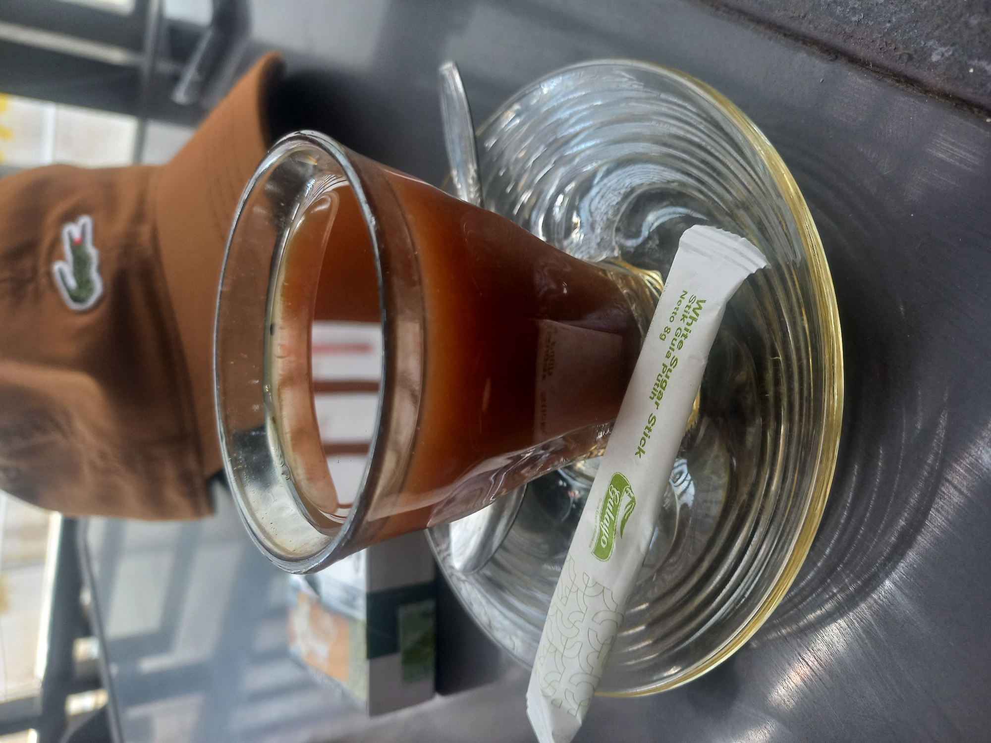 The image presents a close-up, slightly tilted view of a glass of dark reddish-brown beverage, likely tea or coffee, placed on a clear, ribbed glass saucer. A metallic spoon rests within the drink. Alongside the glass on the saucer is a white sugar stick packet, clearly labeled "White Sugar Stick" and "Netto 8g," with additional text "Stik Gula Putih" and a prominent green "Dalego" brand logo.
To the left of the beverage, a brown baseball cap with a distinctive green and white crocodile logo (identifiable as Lacoste) is partially visible, suggesting a casual atmosphere. The entire arrangement is set on a brushed metallic table surface, which shows reflections and a textured grain. The background is softly blurred, indicating other elements or structures within the setting but keeping the focus on the foreground items.
The scene captures a moment of refreshment or leisure, possibly at a café or casual eatery in Titibaro, Indonesia. No specific time of day or weather is discernible, but the lighting suggests an indoor or covered outdoor setting. The image is a factual snapshot of everyday items associated with enjoying a hot drink.