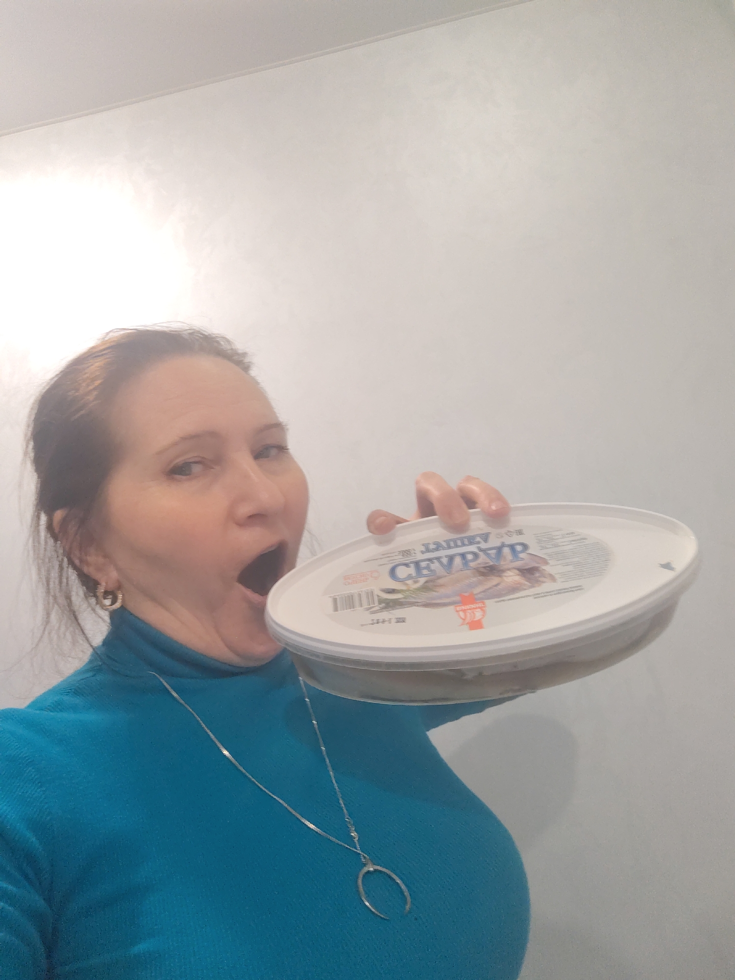 A woman is captured in a playful, mid-shot pose indoors, likely in Sarapul, Russia. She has dark hair pulled back and is wearing a teal turtleneck sweater, a silver necklace with a crescent moon pendant, and silver hoop earrings. Her mouth is wide open in an exaggerated expression, as if about to take a large bite or shouting. She holds a large, round white plastic food container with a lid up to her open mouth, suggesting an act of humorously eating directly from the container.
The container's label is partially visible, prominently featuring the Cyrillic word "СЕВРЮР" (SEVRYUR) and an illustration of fish, likely herring or sprats. Other smaller text, including "ENONNOL," and a barcode are also discernible on the lid. The background is a plain, light-colored wall with subtle texture, indicating an indoor setting under what appears to be artificial light. The image conveys a lighthearted and candid moment.