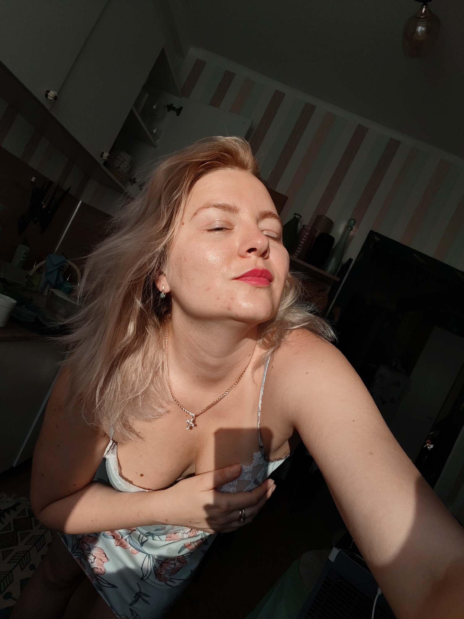 A woman with blonde hair and her eyes closed is seen from the chest up, wearing a light-colored dress with a floral pattern and a necklace with a cross pendant. She is holding her right hand over her chest. The lighting suggests the photo was taken indoors, likely in a kitchen, with sunlight streaming in from the left. The background includes kitchen cabinets, shelves with various objects, and a dark-colored television screen. The environment appears to be a residential setting. The location is Izhevsk, Russia.