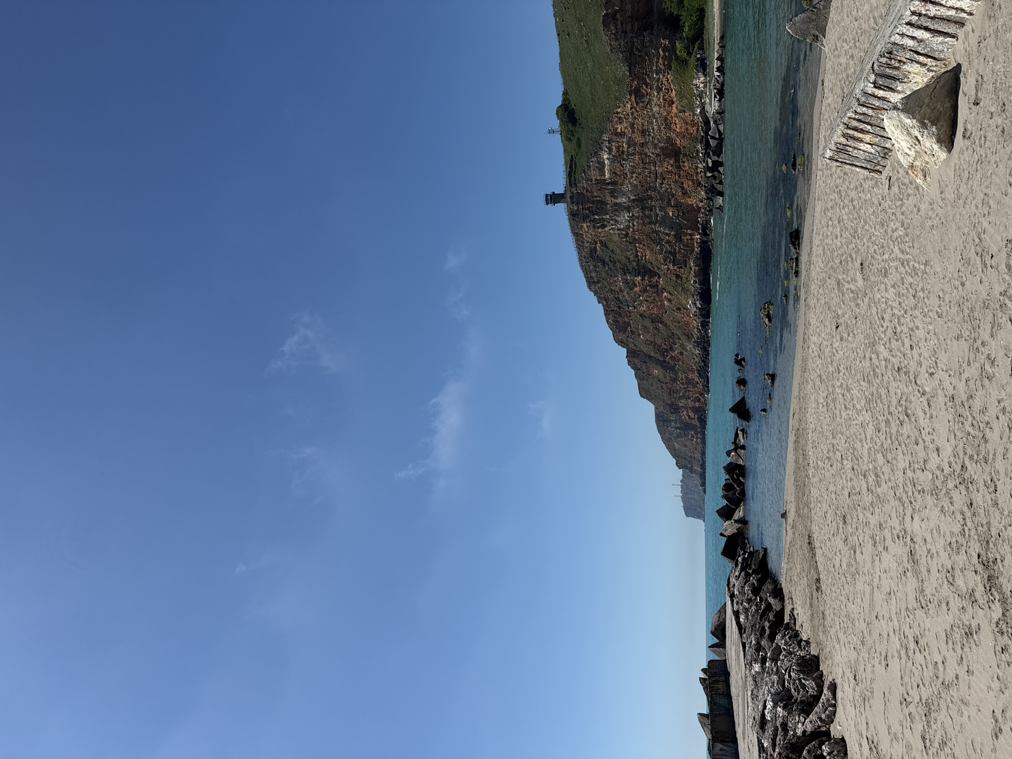 The image depicts a coastal scene near Balgarevo, Bulgaria. A rocky cliff face topped with vegetation occupies the upper right portion of the frame. A tower structure is visible on the cliff. Below the cliff is a narrow beach with light-colored sand. The water is turquoise. A breakwater made of large dark rocks extends into the water along the bottom. Some broken concrete structures are visible on the beach to the right. The sky above is clear and blue, with a few thin clouds.
