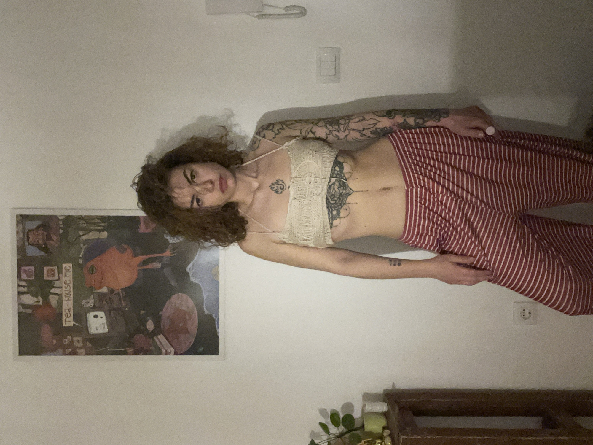 A person is lying against a wall in an indoor setting. The person has curly brown hair and is wearing a light-colored lace top and red and white striped pants. Tattoos are visible on their arms and chest. A framed poster with the words "Tea-house me" is on the wall to the left of the person. A light switch and electrical outlet are also on the wall. Furniture is along the bottom of the wall. The location is inside an unspecified building in Boreti, Montenegro.