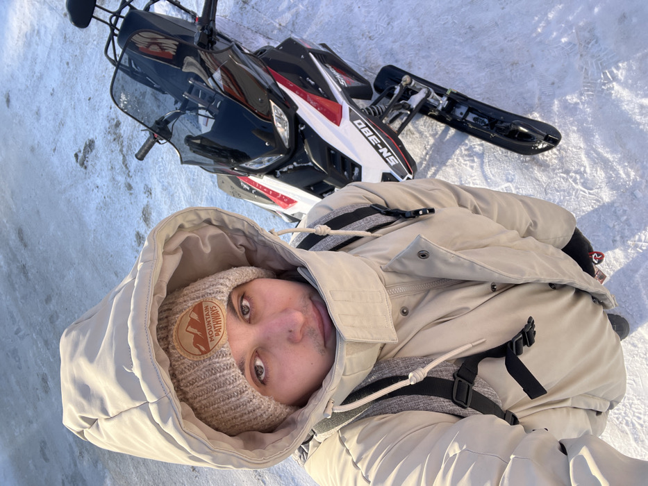 An overhead-angle selfie taken in Васильево, Russia, depicts a male individual standing on snow and ice next to a snowmobile. The man wears a light beige hooded winter coat, a beige knit beanie with an embroidered "MOUNTAIN PATHWAYS" patch, and a dark-strapped backpack visible over his shoulders. He has short facial hair and a small piercing below his lower lip. To his right, a black, white, and red snowmobile is parked, displaying "SN-380" on its side, with its handlebars, windshield, seat, and front skis visible. The ground consists of uneven snow and patches of ice under bright natural light.
