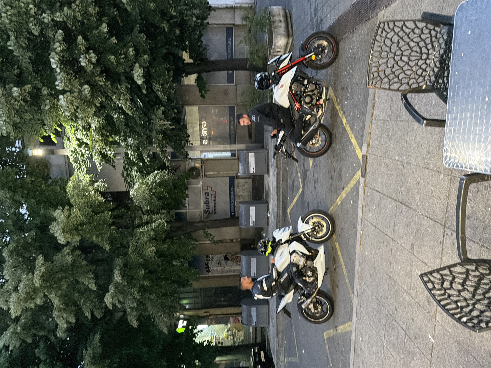 In Burgas, Bulgaria, two men are seated on white motorcycles, parked on a street lined with trees and buildings. The man on the left, wearing a white and black racing suit, sits on a white sportbike. He is looking to his right. A yellow and black helmet rests on the motorcycle's seat. The man on the right, in a black racing suit, is on a white sportbike with red accents and a black helmet. He is looking to his left. Buildings with shopfronts are visible in the background, including one with "Subra" signage. Two large grey trash bins are positioned on the sidewalk. A black car is parked to the left of the frame. In the foreground, a black metal chair and table are visible, suggesting an outdoor seating area. The lighting indicates it is either early morning or late evening.