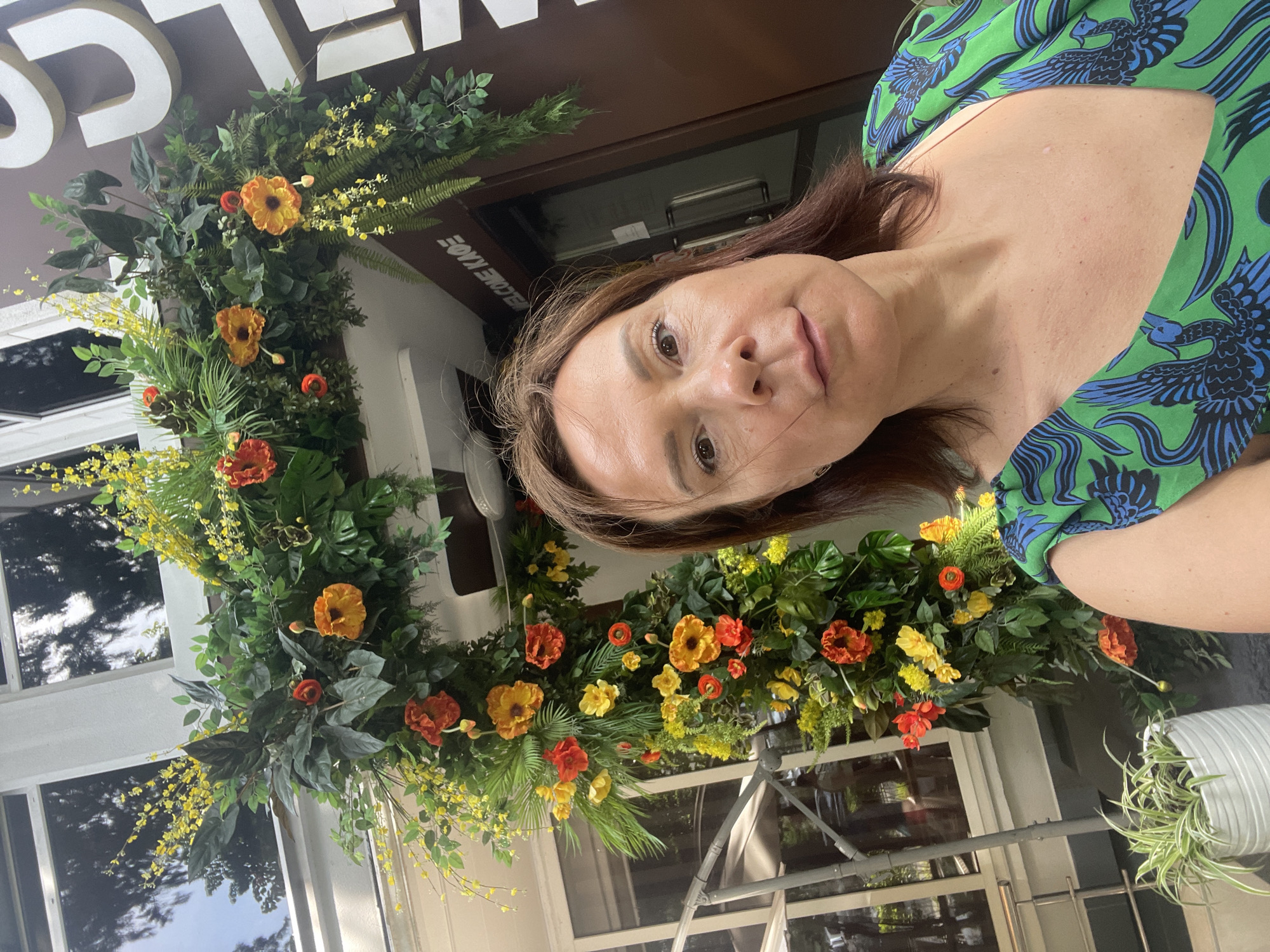 A medium-shot, eye-level selfie shows a woman in the foreground, facing the camera with a neutral expression. She has brown hair and is wearing a green dress with a blue bird print. Behind her, an elaborate floral archway frames an entrance. The archway is adorned with numerous artificial flowers in shades of orange, red, and yellow, along with lush green foliage. The entrance behind the archway has a dark brown door and a sign with white text reading "WELCOME KAOE". To the left of the archway, a white potted plant with long, green leaves is visible. The overall scene appears to be outdoors, possibly in front of a cafe or establishment, in the city of Sochi, Russia.