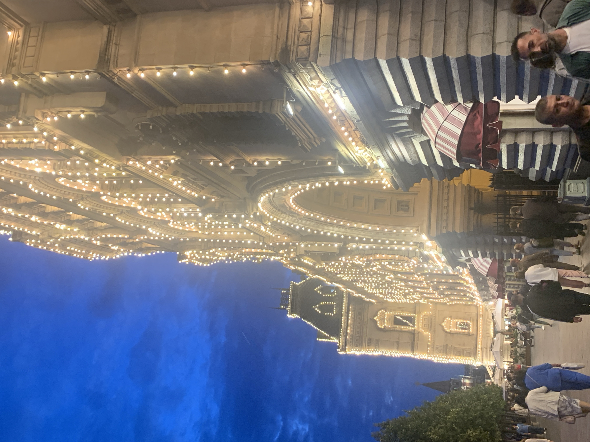 The image depicts a nighttime scene on Red Square in Moscow, Russia, likely during an evening when the buildings are illuminated. The central focus is the facade of a grand, historic building, possibly the GUM department store, heavily adorned with strings of warm, white lights that trace its architectural details, including arched windows and cornices. To the left, another building with a steeply pitched roof, also outlined in lights, is visible. The sky above is a deep twilight blue with some wispy clouds.
In the foreground and mid-ground, a crowd of people are walking along a pedestrian area. The individuals are dressed in casual, everyday clothing. Some are walking in pairs or small groups, suggesting they are tourists or locals enjoying the evening. The atmosphere appears lively and inviting, typical of a popular public space. There are trees lining the walkway, and in the distance, umbrellas of outdoor seating areas for cafes or restaurants are visible. The overall impression is one of a bustling urban environment at dusk, enhanced by festive illumination.