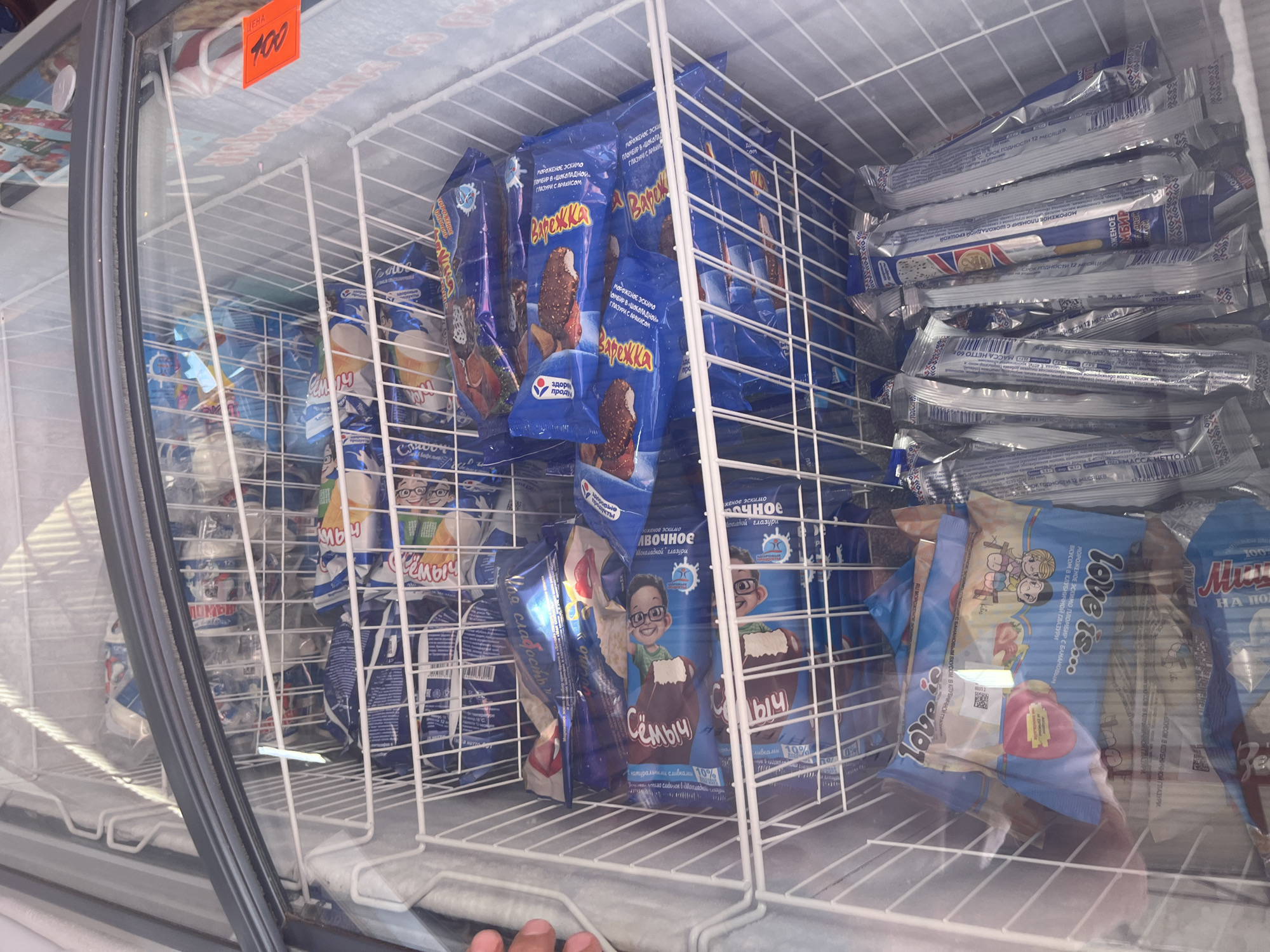The image shows an open freezer chest filled with packaged ice cream. The freezer is divided into shelves by white wire racks. The ice cream packages are primarily blue and white, with some showing images of ice cream cones, cartoon characters, and text in Cyrillic. Some of the packages have orange price tags attached. The freezer is located inside a store or market in Plastunovskaya, Russia, as indicated by the price tag written in Russian. The lighting suggests it is daytime, with natural light filtering into the freezer. There are no people visible in the image, and the focus is solely on the frozen goods. The scene is a typical retail display of frozen desserts.
