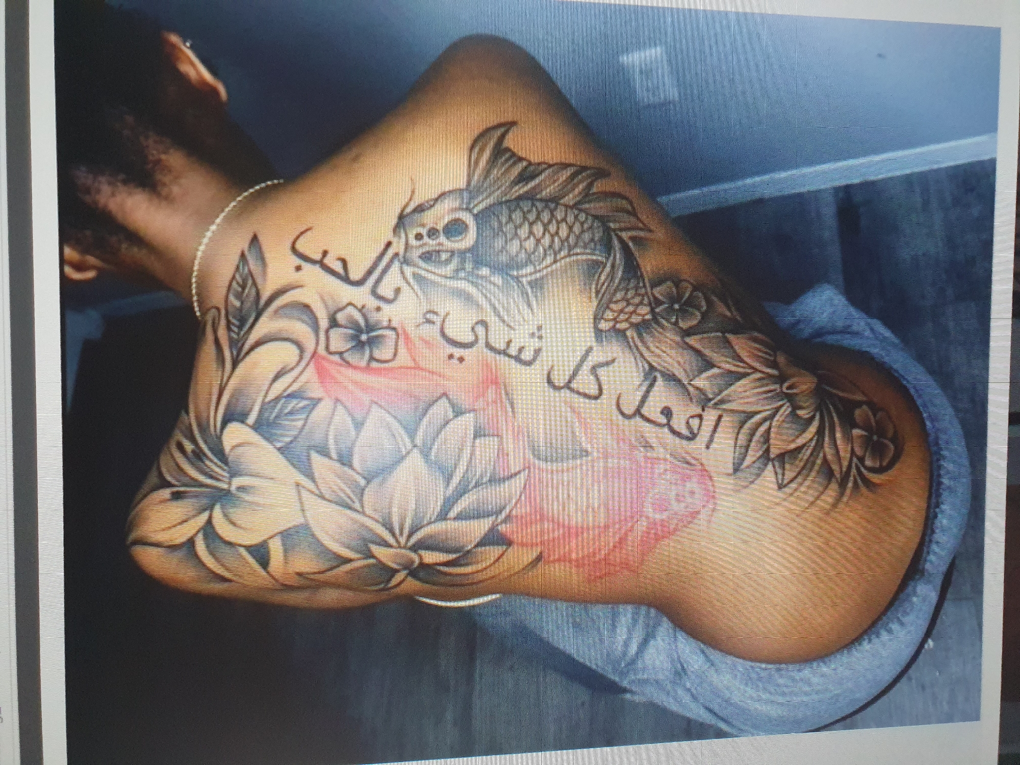 The image shows a person's shoulder and upper arm, tattooed with a design featuring a koi fish, lotus flowers, and Arabic script. The tattoo is rendered in black and grey, with some subtle pink and red shading in the lotus flowers. The koi fish is depicted in the center of the tattoo, with its body curving around the arm. The lotus flowers are arranged around the fish, with some open and others closed. The Arabic script is positioned above and below the fish, woven into the floral elements. The skin tone of the person is dark, and they are wearing a light-colored bracelet on their wrist. The background is dark, with hints of a wall and possibly a curtain or garment. The lighting is somewhat dim, highlighting the tattoo. There are no clear indicators of time of day, weather, or specific location within Tashkent, Uzbekistan, beyond what can be inferred from general indoor lighting. The emotion conveyed is difficult to determine solely from the image, but the intricate and colorful tattoo suggests a personal and artistic expression.