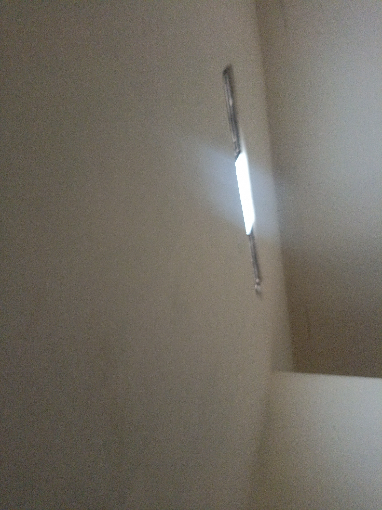 The image shows a light fixture mounted on a plain, light-colored wall. The light fixture is elongated and appears to be emitting a bright white light. The wall has a textured appearance. The overall scene is indoors and dimly lit, with the light fixture being the primary source of illumination. The image does not provide any specific information about the city of Peganden, Indonesia, nor does it depict any people, actions, or interactions. It is a static environmental shot focused on the light fixture and the wall.