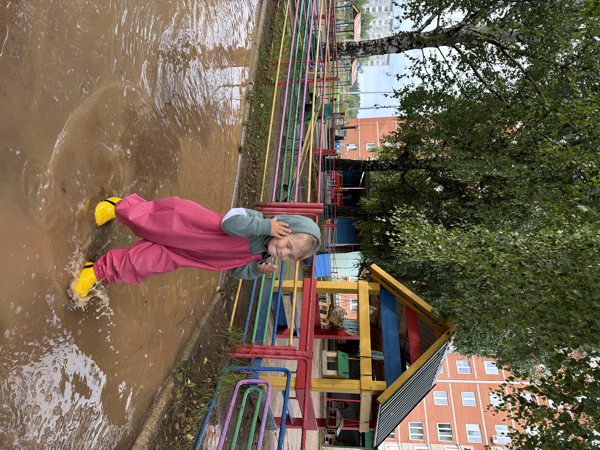 A young child wearing pink overalls and yellow boots splashes in a muddy puddle on a playground in Chaikovsky, Russia. The child is looking to the right with their hands near their hood. In the background, there is a colorful wooden playhouse, fences, trees, and apartment buildings. The sky is overcast.