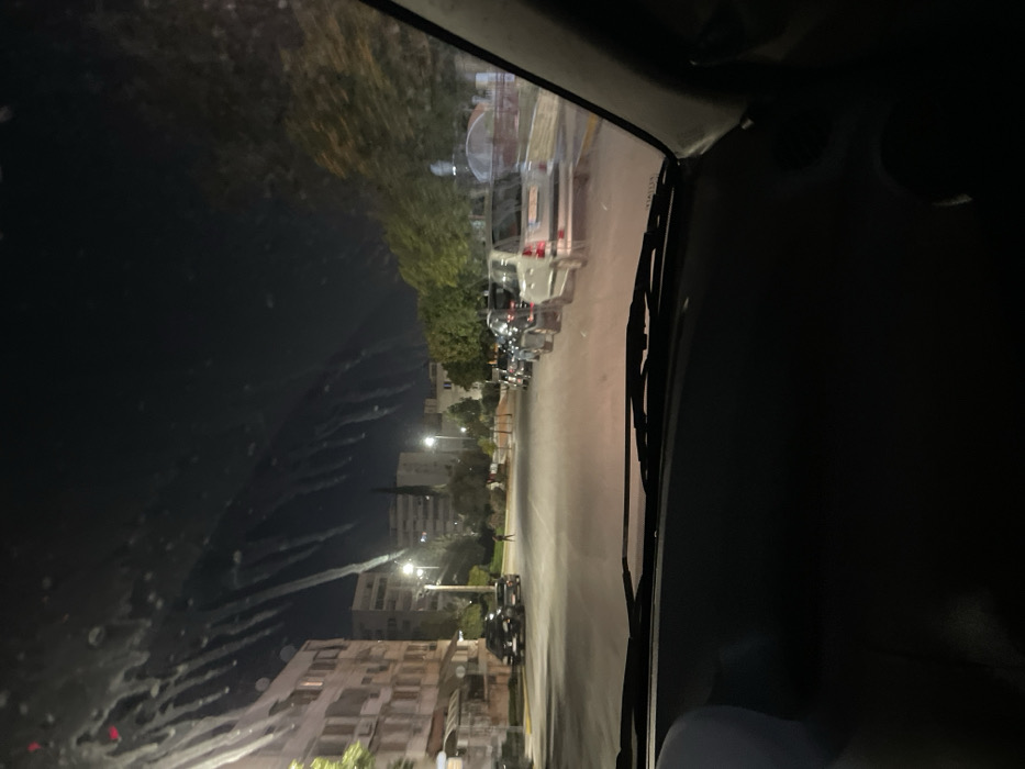 This night-time image, taken from within a vehicle in Drapetsona, Greece, looks out through a streaked windshield, possibly affected by rain or dirt. The sky is dark, indicating late evening or night.
In the immediate foreground, parts of the vehicle's interior, including a dashboard area and a prominent windshield wiper blade, are visible on the right side. The perspective is from the driver's or front passenger's seat.
Beyond the windshield, a street scene unfolds. Cars are parked along the left side of the road, including several white vehicles and darker cars, suggesting a residential or urban environment. Multi-story buildings, typical of European architecture, line both sides of the street. Interspersed among the buildings and alongside the road are trees with green foliage. Artificial lighting from streetlights illuminates the scene, casting a warm glow on the road and surrounding structures. Further down the street on the left, a small, dark figure, likely a pedestrian, is faintly visible.
The background shows more buildings under a dark sky, which exhibits some faint, wavy streaks that could be reflections on the glass or subtle cloud formations. The overall activity appears limited to a quiet street scene at night, with no discernable dynamic events. No clear, readable text is visible on any objects or signs within the frame. The atmosphere is still and calm, characteristic of an ordinary night in a town.