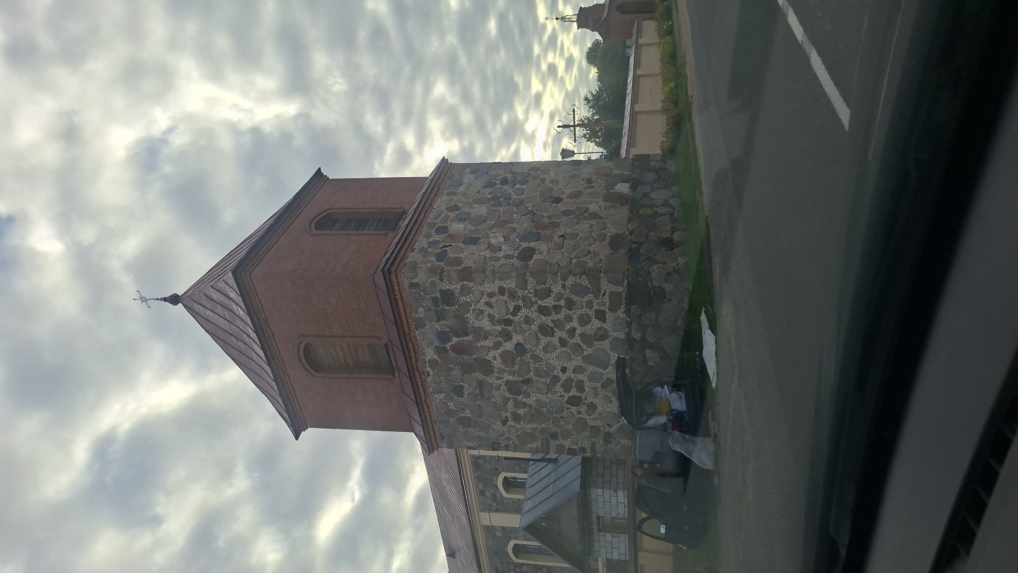 The image captures a view of a church tower and adjacent building in the Валкалацкі сельскі Савет, Belarus. The church tower, constructed from large, irregularly shaped stones at its base and red brick on its upper section, is topped with a copper-colored, pyramid-shaped roof and a cross. To the left of the tower, a portion of the church's main building is visible, featuring a red roof and arched windows. In front of the church's base, a dark-colored car with its trunk open is parked on a grassy area next to a road. A person can be seen sitting in the driver's seat, and various items are piled in and around the open trunk, including what appears to be bags and possibly a white sheet on the ground. The sky is overcast with dramatic cloud formations. A fence runs along the side of the road, and further in the distance, another smaller church structure or bell tower is visible. The overall scene suggests a potentially overcast day with some activity around the parked vehicle at the church location.