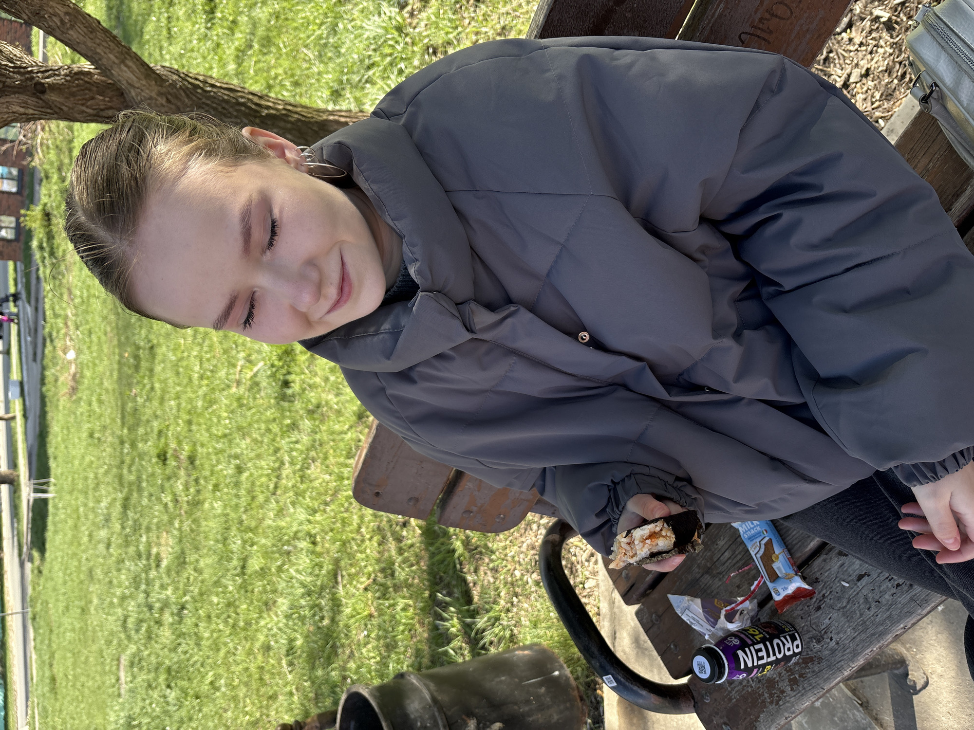 A young woman sits on a park bench outdoors in Chaikovsky, Russia, eating sushi. She wears a grey puffer jacket and dark pants. Her hair is pulled back. She holds a piece of sushi in her left hand. Discarded wrappers and a "Protein" branded drink can are on the bench next to her. Green grass and a tree are visible in the background.
