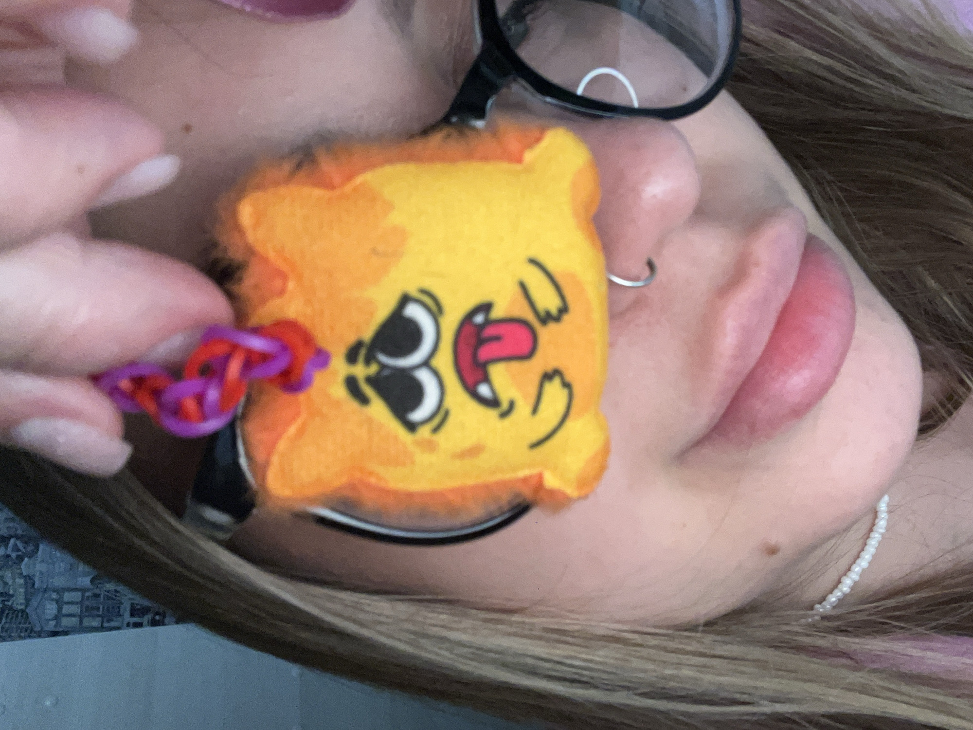 A close-up, slightly tilted image shows a person holding a yellow, square-shaped plush toy/keychain with cartoonish face details. The keychain is attached to the person's eyeglasses. The person's face is partially visible, including their lips, nose, and part of an ear with a beaded earring. The person has brown and blonde hair, and pink lipstick. A section of what may be a loom-band is visible near the keychain. The environment is unclear, but what can be made out suggests an indoor setting. The location Novopokrovskaya, Russia is not apparent within the image.