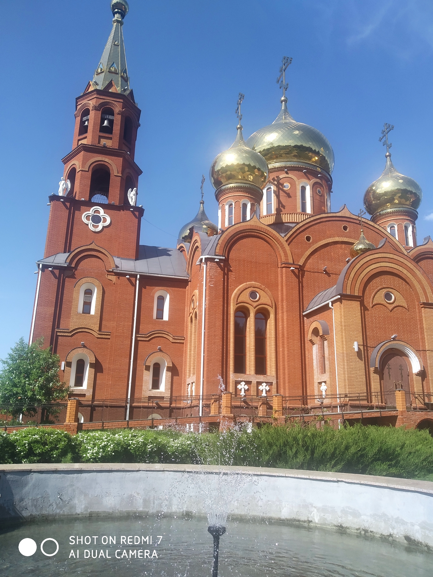 The image shows a red brick Orthodox church with golden domes under a clear blue sky. The church has a tall bell tower to the left, topped with a green spire. The lower part of the image shows a fountain with water spraying. The church is located in Chaikovsky, Russia. Green shrubbery and small white flowers are in front of the church. The fountain is in the foreground. The text "SHOT ON REDMI 7 AI DUAL CAMERA" is visible at the bottom.
