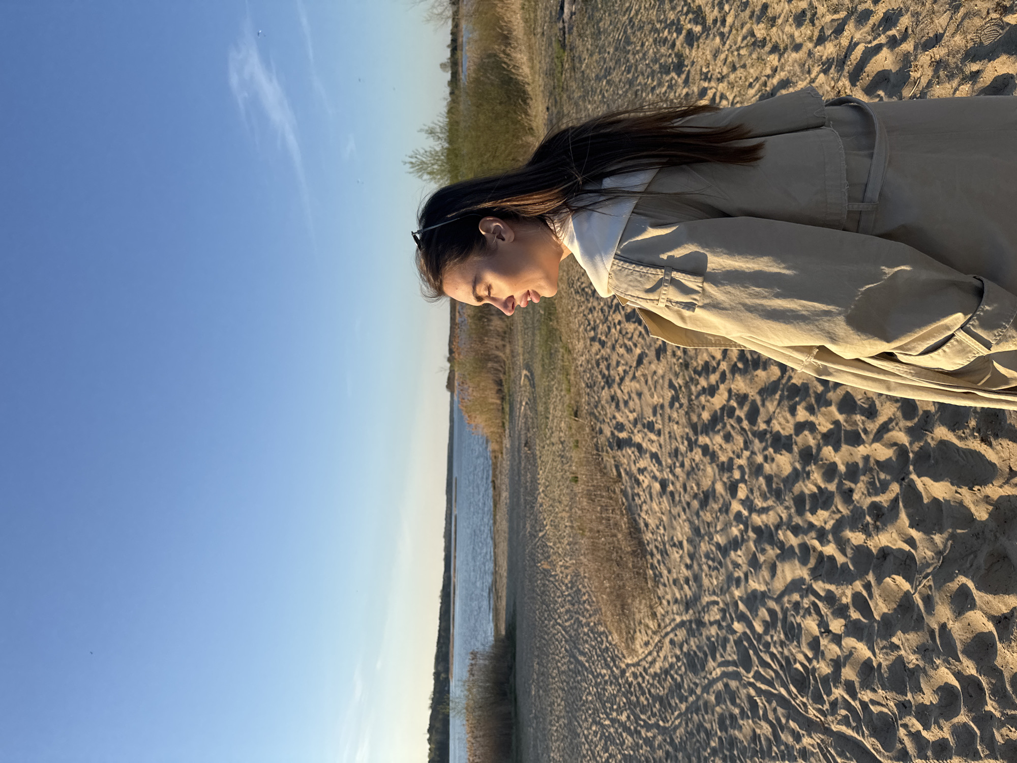 A young woman stands on a sandy beach, likely in Arciechów, Poland. She is looking down and wearing a beige trench coat over a white hoodie. Her dark hair is long and straight. Sunglasses are perched on her head. The beach is bordered by a body of water, possibly a lake or river, with reeds growing along the edge. The sky is blue and clear. The scene is bathed in golden sunlight, suggesting it is either early morning or late afternoon.