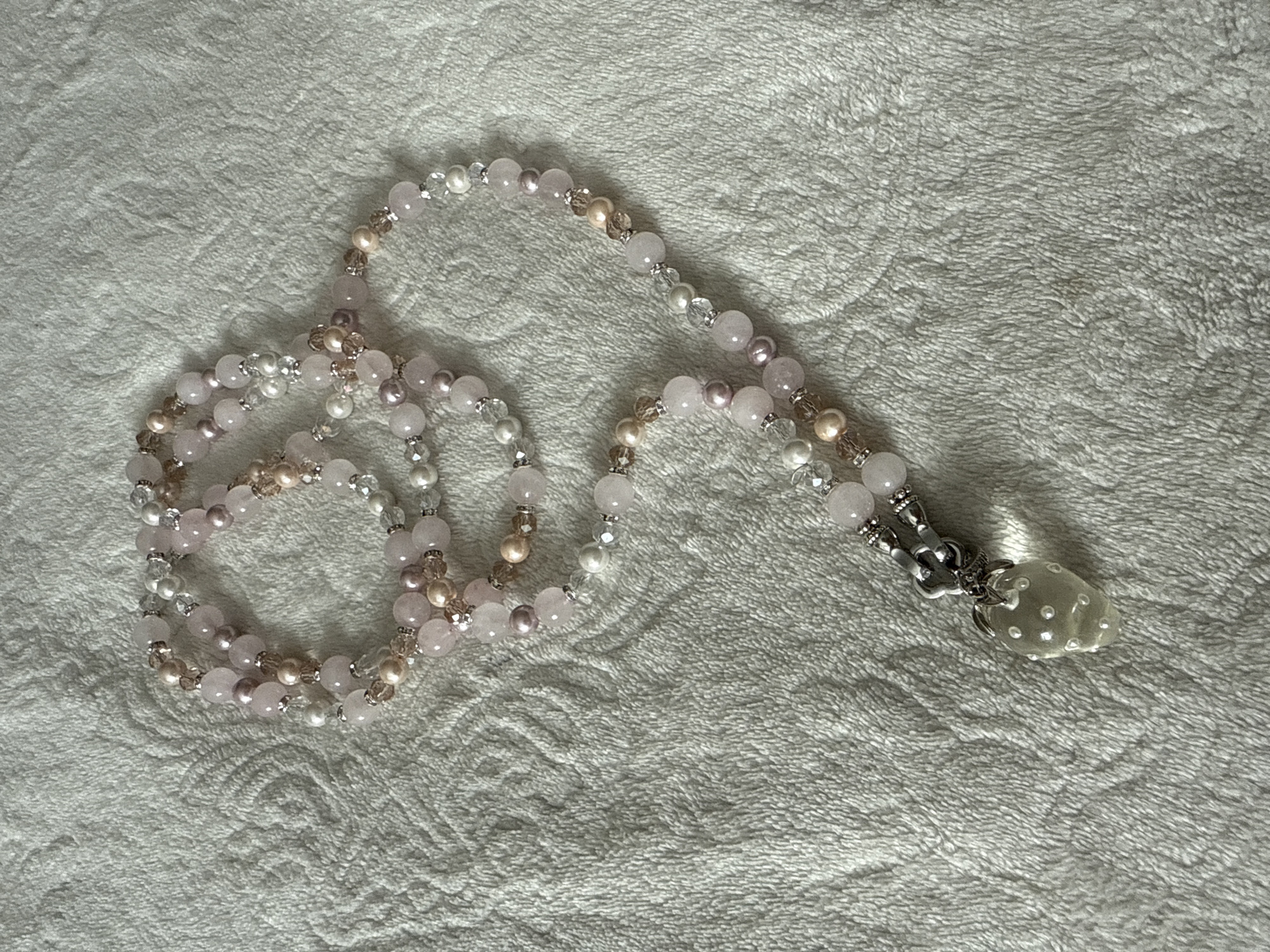 A beaded necklace with a strawberry charm lays on a textured white surface. The necklace is made up of pink, white, and iridescent beads of various sizes. The strawberry charm is clear and appears to be made of glass, with tiny white beads embedded in it. The background is a soft, fuzzy white material with a subtle embossed pattern. There are no people or animals present in the image.