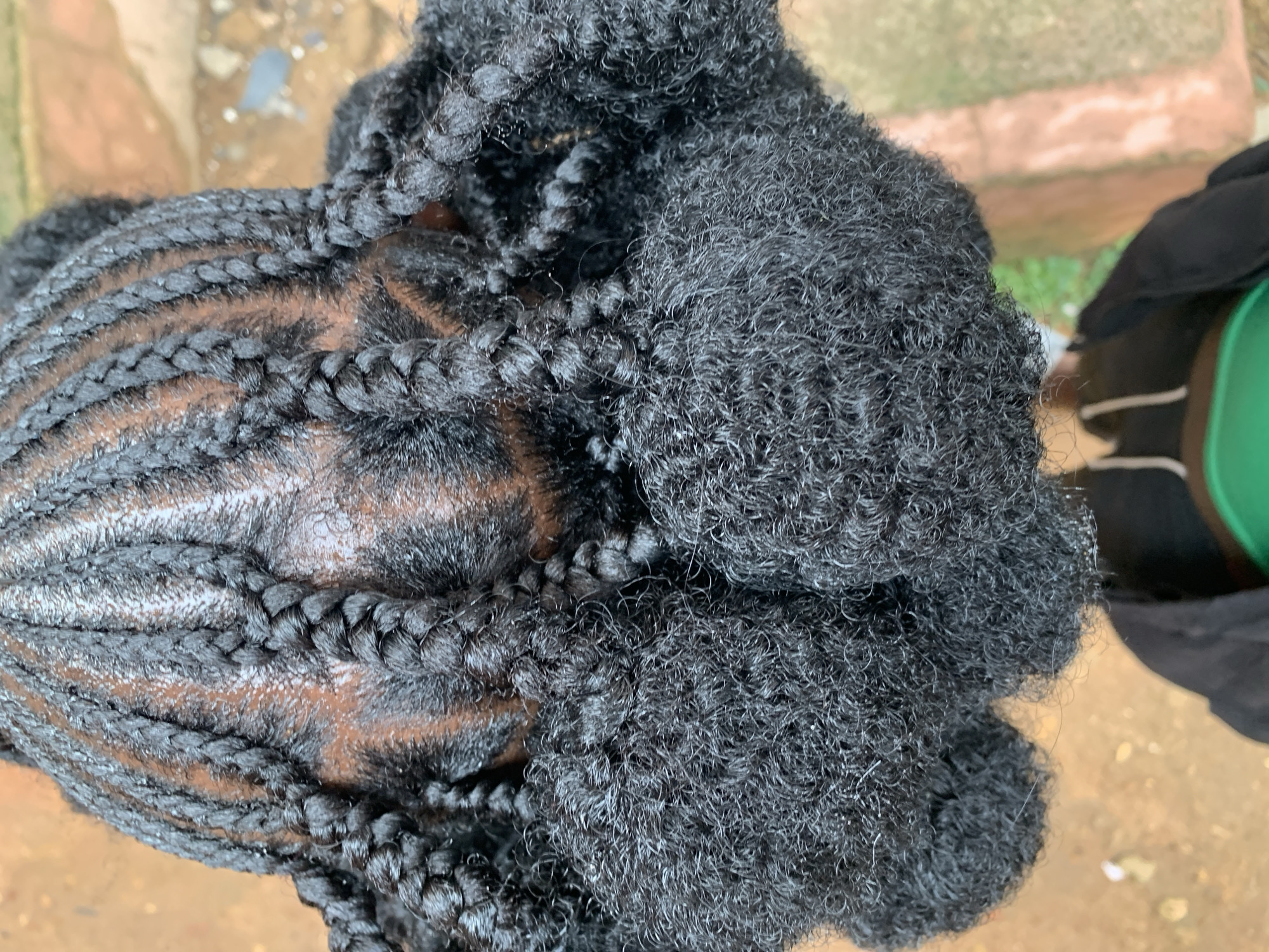An overhead, close-up shot shows the back of a person's head with their hair styled in cornrows leading to large, fluffy buns. The cornrows are neat and tightly braided, parting the scalp in a symmetrical pattern. The buns are voluminous and made of dark, textured hair, creating a striking contrast with the smooth braids. The background appears to be an outdoor setting, possibly in New Stadium, Nigeria, with hints of a reddish-brown, earthy surface, suggesting a dusty or unpaved area, and some patches of green foliage. The lighting is natural and diffused.