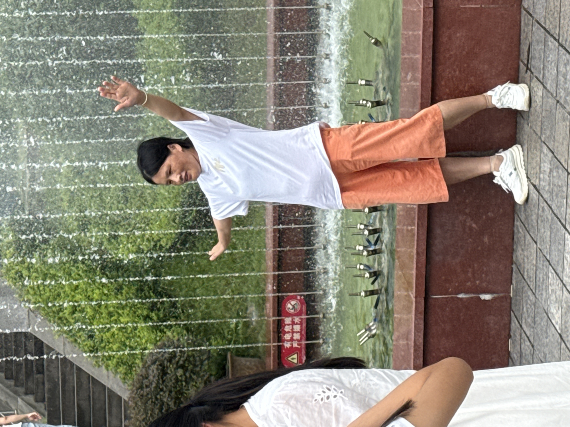 A woman is performing Tai Chi in front of a fountain in Yuzhong District, China. The woman is wearing a white t-shirt and orange shorts. She is in a dynamic pose with her arms outstretched. The fountain is spraying water, and there are green bushes behind it. A red sign with Chinese characters is visible on the edge of the fountain. The ground is paved with square tiles. A portion of another person, wearing a white dress, is visible on the left side of the image. Steps are visible in the background on the left.