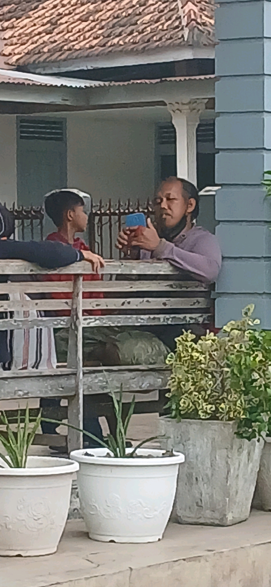 A man with a beard and dark hair is seated on a wooden bench, holding a blue cell phone. He is looking at the phone, and his face is illuminated by its light. Next to him, a young boy wearing a red shirt and a hat is looking at the man. They appear to be interacting. The bench is located on a porch or veranda, with a tiled roof and a white column visible in the background. There are also potted plants in front of the bench. The image was taken in the city of Metro, Indonesia.