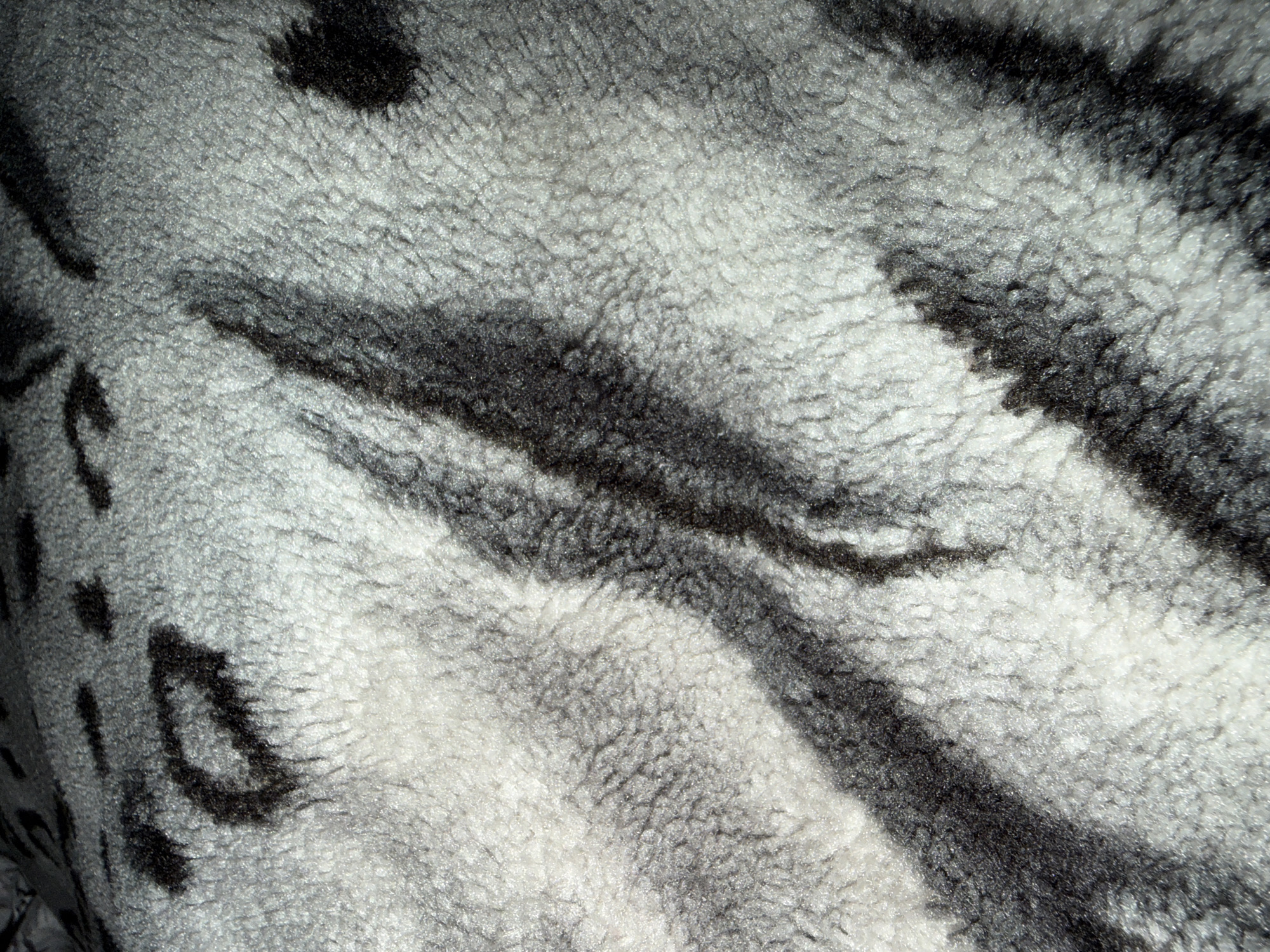 This is a close-up, macro photograph of a textured surface, likely a fabric or animal pelt, with a pattern resembling that of a snow leopard or a similar spotted big cat. The texture is fluffy and soft, with visible individual fibers creating a sense of depth and materiality. The pattern consists of irregular, dark grey to black splotches and stripes against a lighter, off-white or pale grey background. The lighting creates highlights and shadows, emphasizing the three-dimensional quality of the surface. The overall impression is one of natural camouflage and tactile softness. There are no people, specific objects, or a discernible scene or setting visible, as the image is entirely focused on the abstract pattern and texture of the material. No text is present. Given the location context of Beșalma, Moldova, this could be a piece of clothing, upholstery, or a taxidermied item, though the specific context within Beșalma is not visually represented.