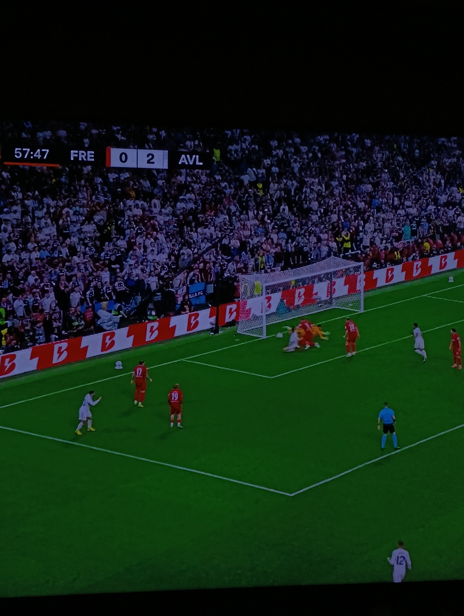 The image shows a soccer match in progress. The score is FRE 0, AVL 2 at 57:47. The field shows a goalmouth scene with players clustered around the net. The majority of spectators in the background are wearing white. The location and city of Plovdiv, Bulgaria are not readily apparent from the image, and no supporting information suggests that these elements are evident.