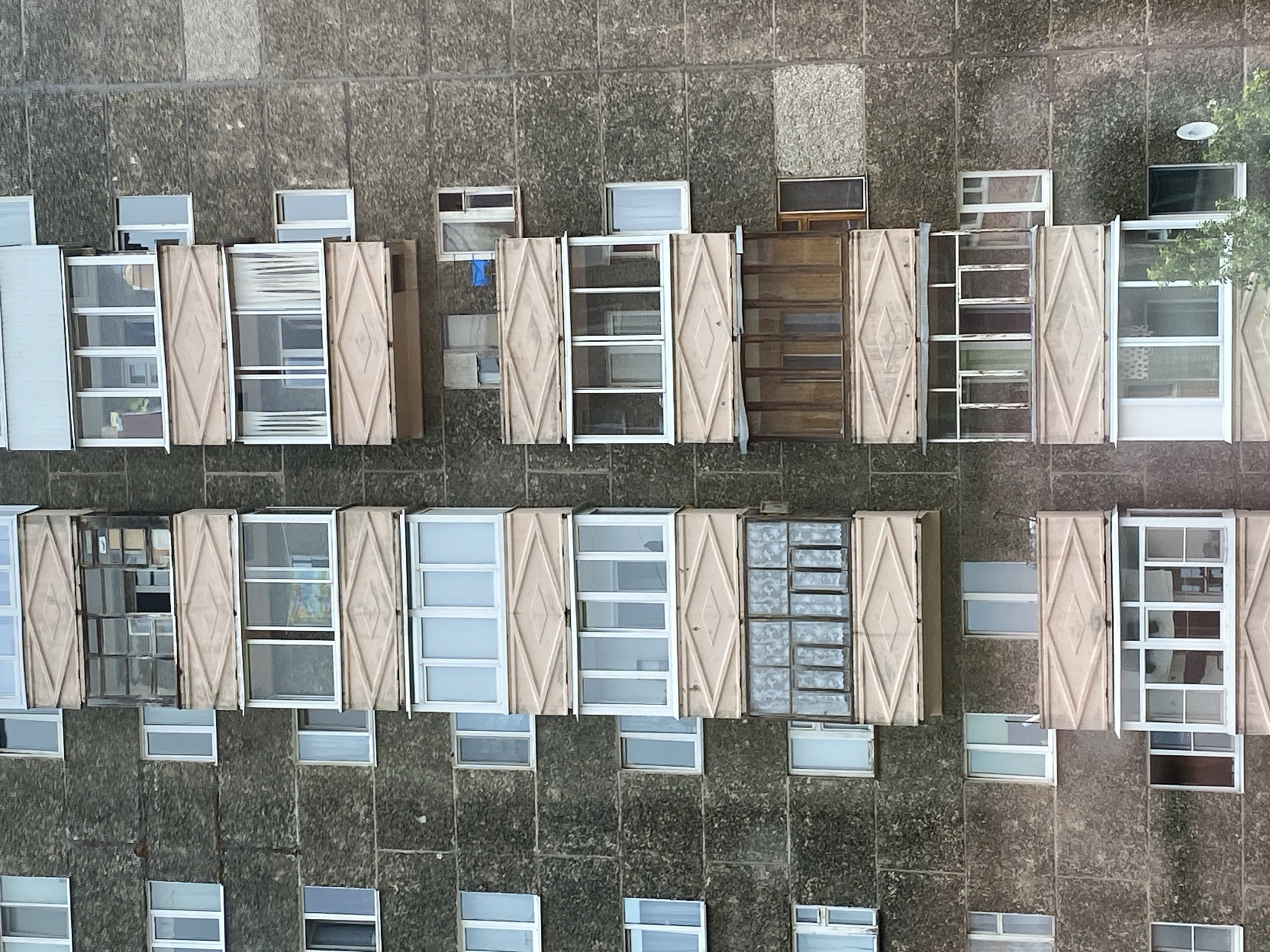 The image shows a section of a weathered, grey concrete apartment building in Votkinsk, Russia. The building features multiple floors with numerous windows and balconies. The balconies are varied in design, some enclosed with glass and others open. Several balconies have decorative pink, diamond-patterned panels. The windows are mostly rectangular and appear to be made of white frames. Some windows have curtains visible. The concrete exterior exhibits signs of age and wear. The sky is not visible in the frame. There are no people or any discernible activity in the image. The overall impression is of a typical, functional residential building in a Russian urban environment.