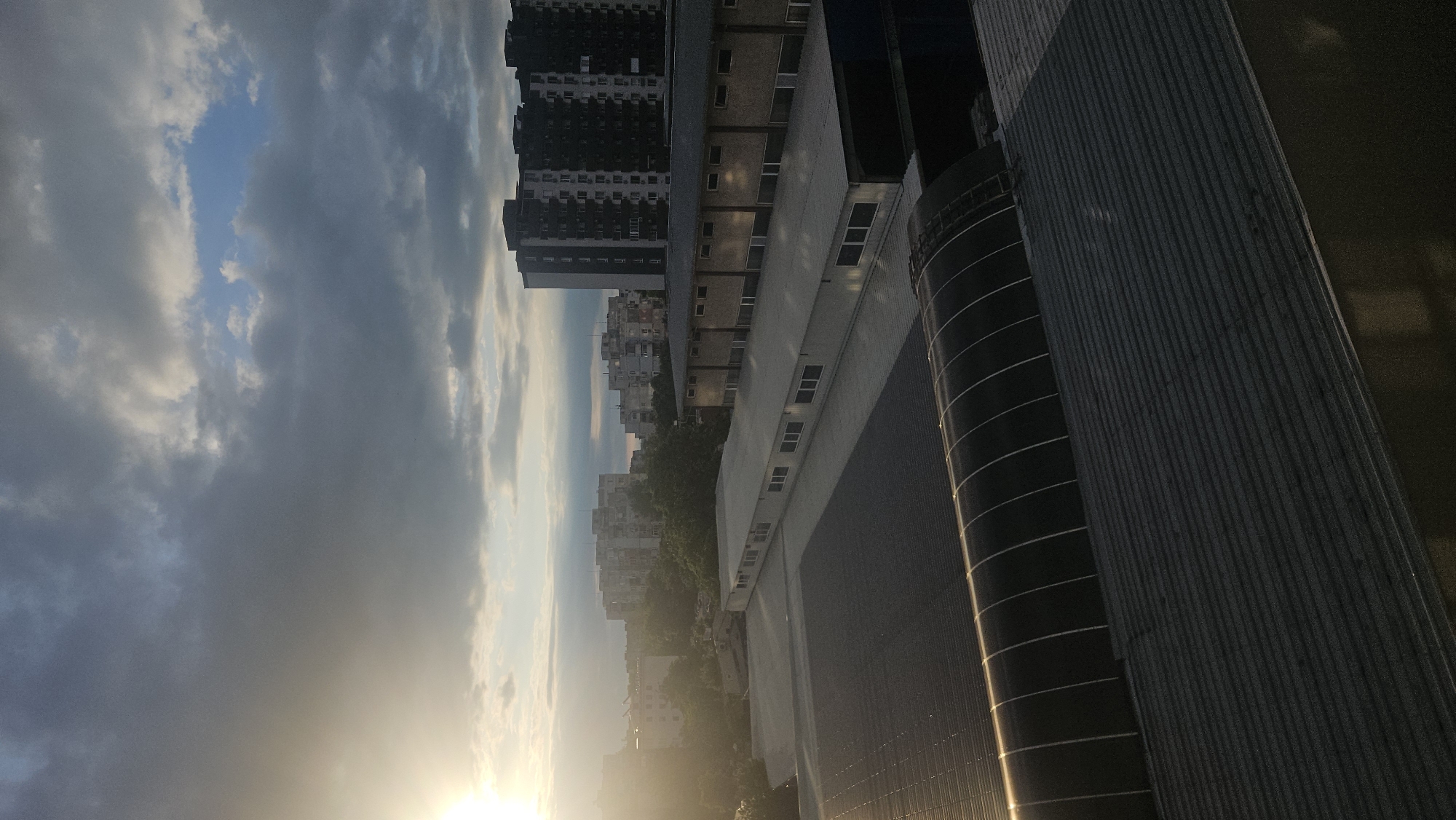 The image shows a view of Plovdiv, Bulgaria, at sunset or sunrise. In the foreground are portions of buildings, including a corrugated metal structure and a cylindrical tower-like feature with horizontal bands. In the midground, there are several apartment buildings and some greenery. The background consists of a sky filled with clouds. The light in the image suggests the sun is either rising or setting, casting a golden glow on parts of the scene. The image is rotated approximately 90 degrees counter-clockwise.