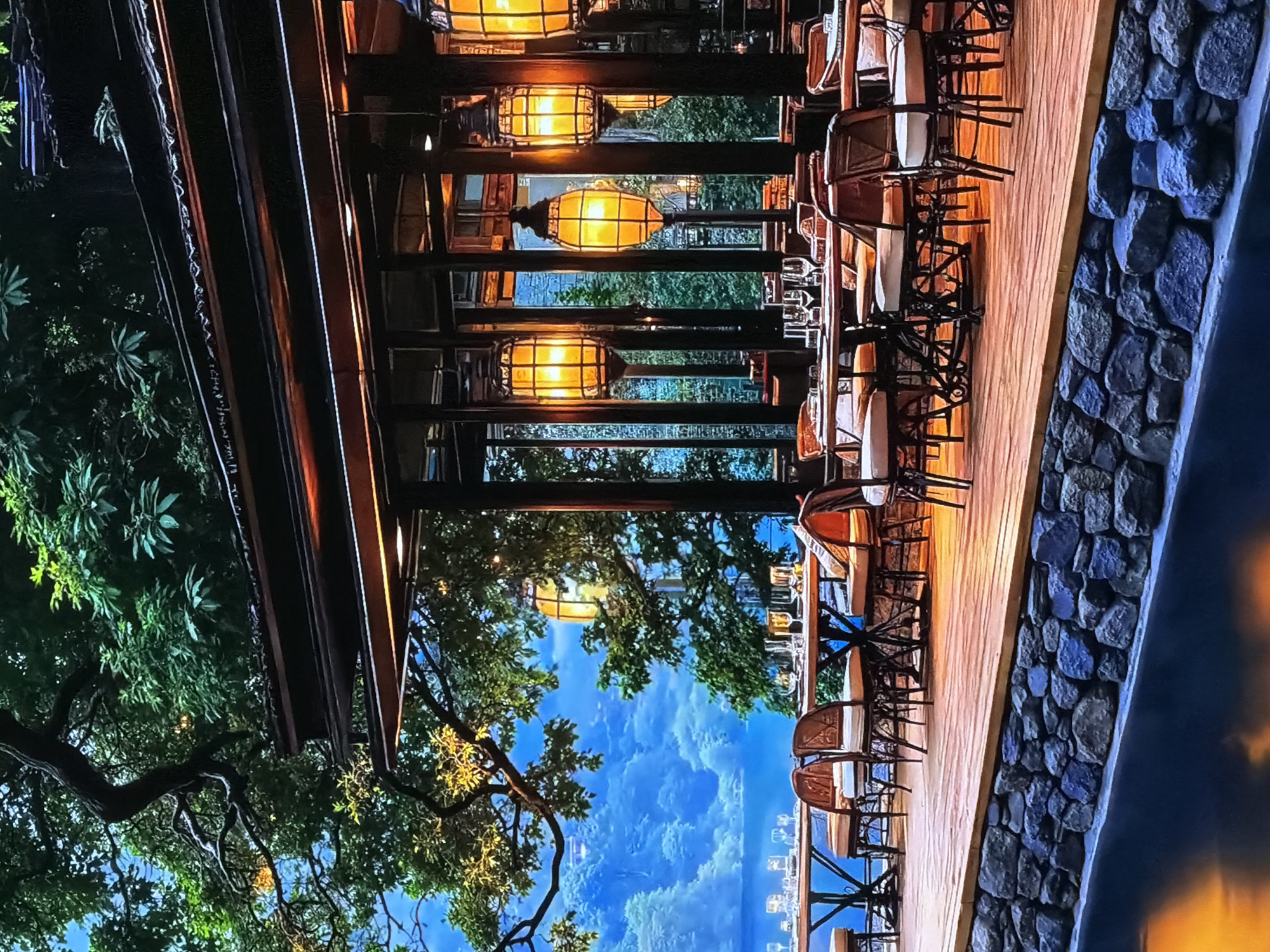 The image captures a serene outdoor dining or lounge area, likely a restaurant terrace, set against a panoramic natural backdrop. The scene is viewed at what appears to be dusk or pre-dawn, illuminated by several warm, golden lanterns with a distinctive cage-like design, hanging from a dark wooden ceiling structure. Vertical wooden beams support the roof, and the floor is composed of light-colored wooden planks.
Along the right side, multiple tables are arranged with chairs featuring woven rattan backs and light-colored cushions, set for guests. Various glassware, including wine glasses, are visible on the tables. A rustic stone wall forms a barrier along the edge of the terrace, beyond which a dark, reflective surface, possibly water, is visible.
To the left, dense green foliage and tree branches frame the view, emphasizing the natural environment. In the distance, a sprawling landscape of rolling hills or forests extends under a blue-hued sky, which could be twilight or early morning. The distant scenery appears soft and hazy, creating a sense of depth.
The setting exudes a tranquil and elegant ambiance, blending natural materials like wood and stone with refined furnishings. There are no people visible, and no discernible text. This inviting outdoor space, offering expansive views and a comfortable atmosphere, is located in Sofiivska Borshchahivka, Ukraine.